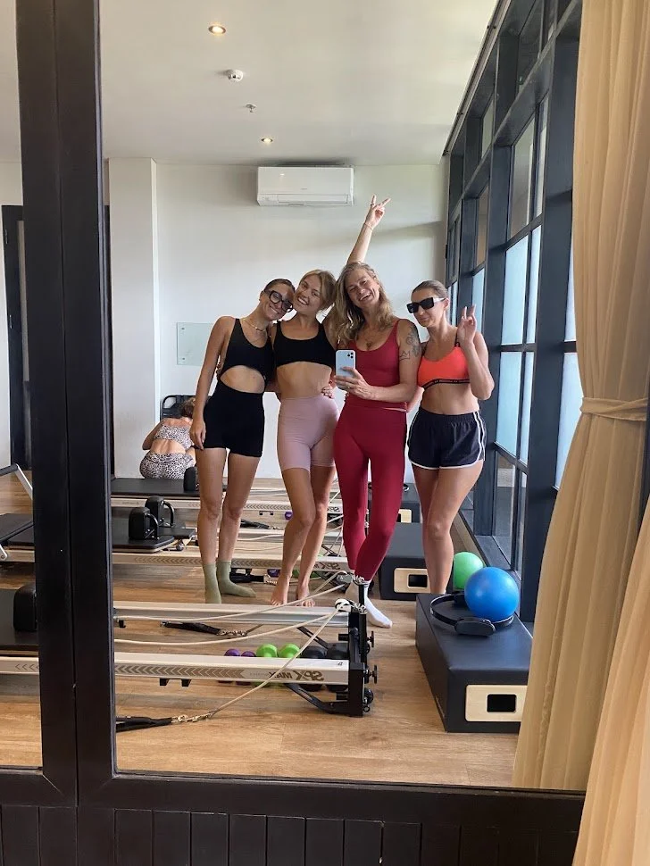 Four women are standing in a fitness studio taking a selfie in a mirror. They are dressed in workout clothes and making peace signs. Behind them, there are exercise equipment and colorful balls, with a large window on the right side.