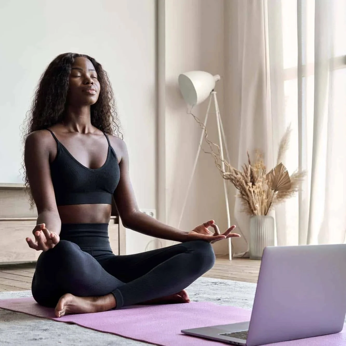A woman practicing yoga indoors, sitting in a cross-legged meditation pose on a pink yoga mat, with her eyes closed and hands resting on her knees.