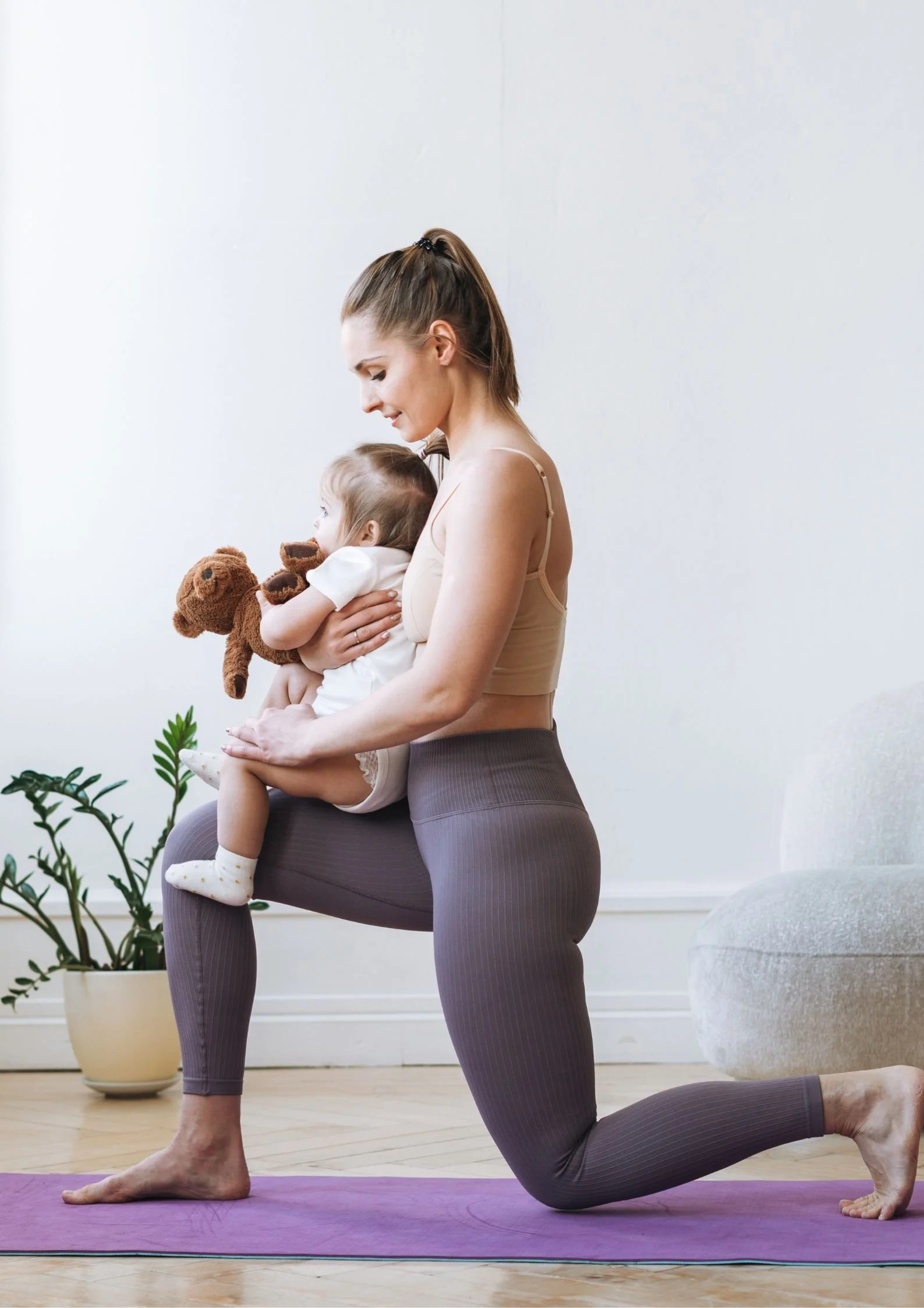 A woman in workout clothes practicing yoga on a purple mat while holding a young girl with a teddy bear in her arms in a bright living room with a potted plant and a white couch.