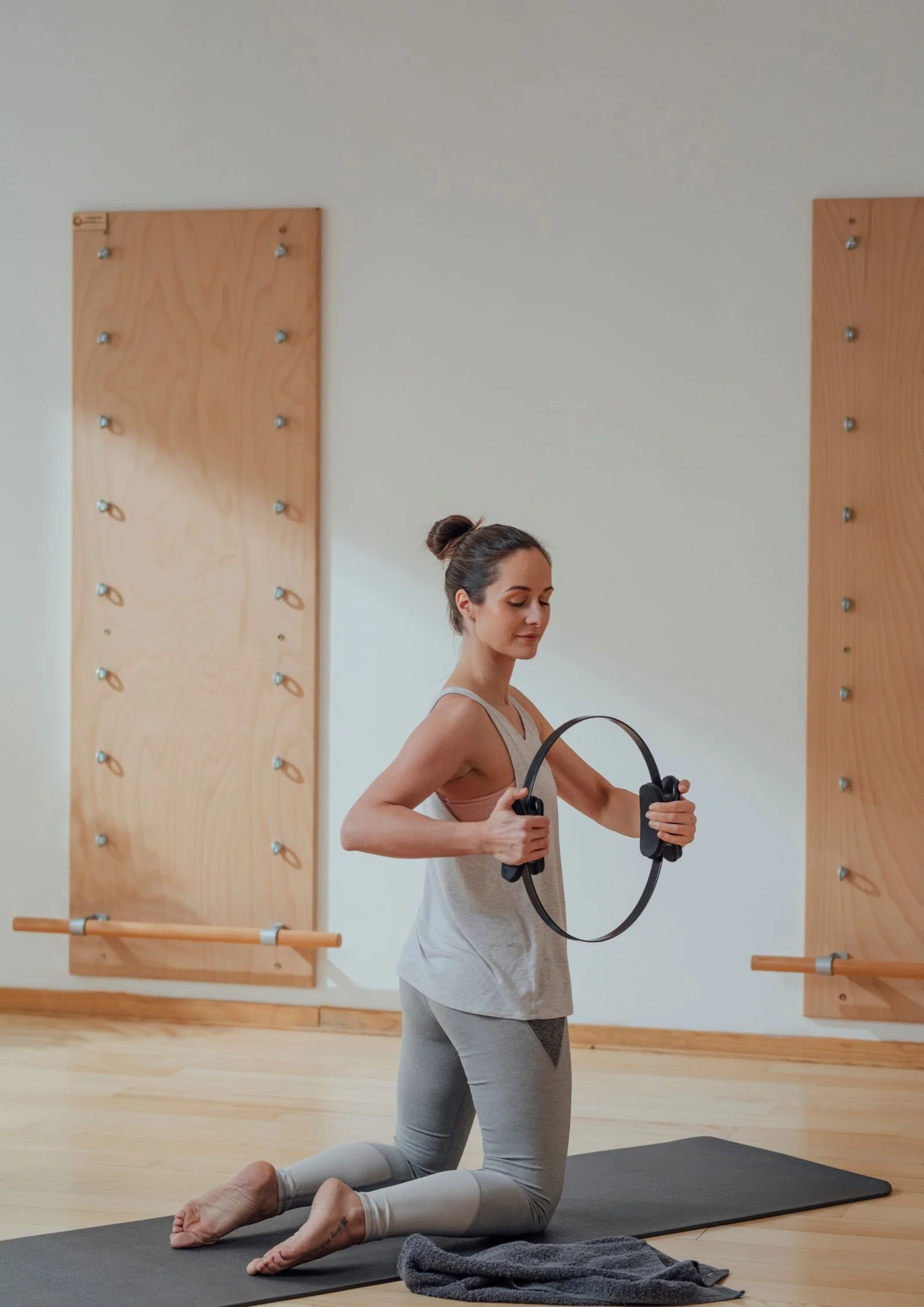 Woman practicing yoga on a mat in a studio, holding a circular fitness ring while kneeling on her knees with a towel nearby.