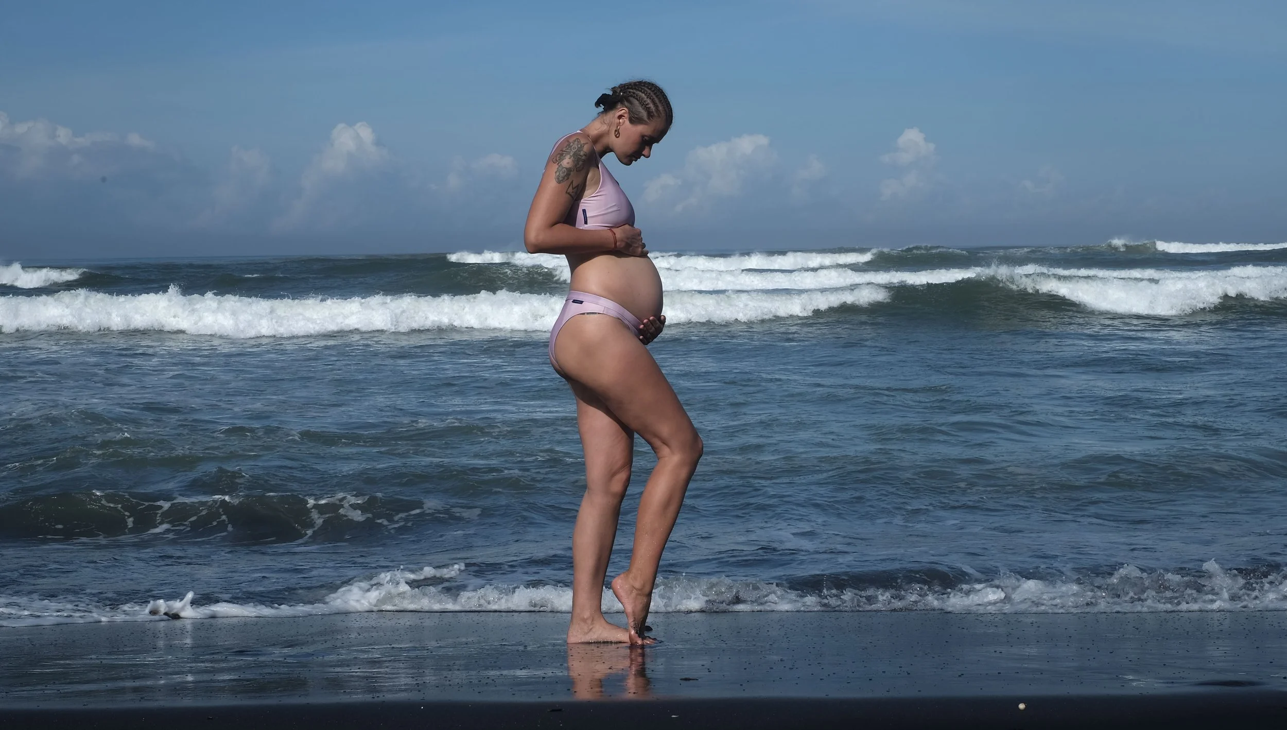 A pregnant woman standing at the edge of the ocean, looking down at her belly while standing in the shallow water with waves rolling in behind her in a beach setting.