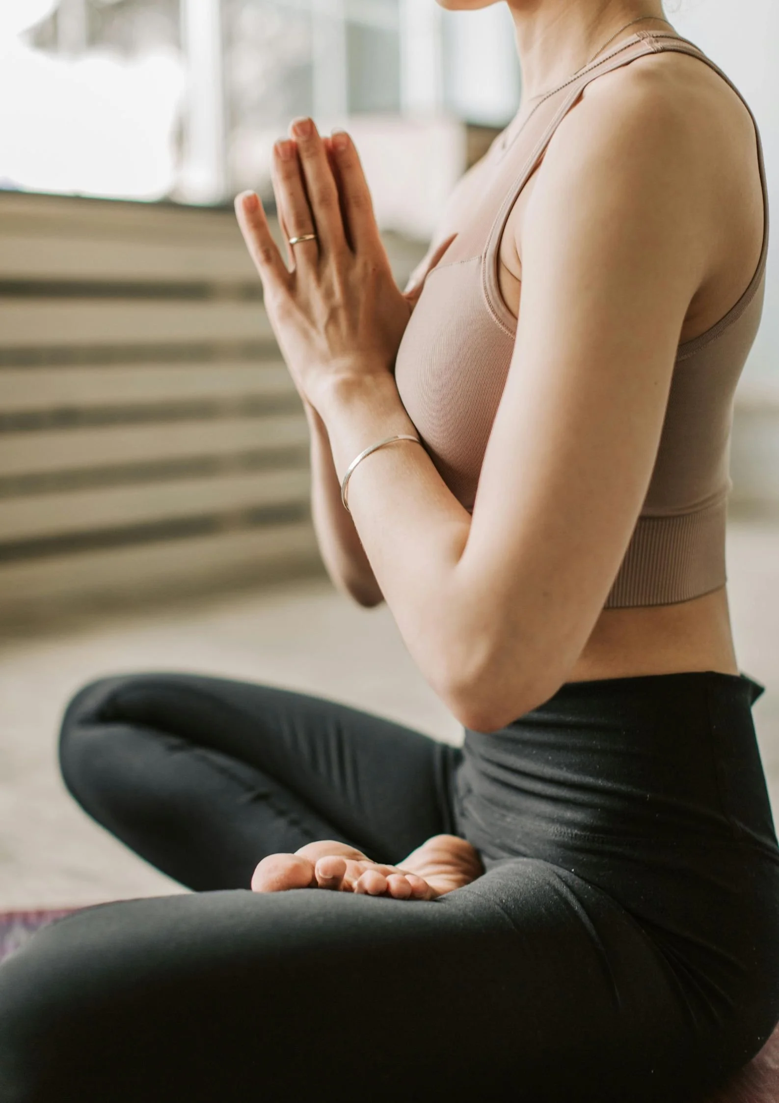 A woman practicing yoga indoors, sitting cross-legged with hands in prayer position, wearing a beige sports bra and black leggings.