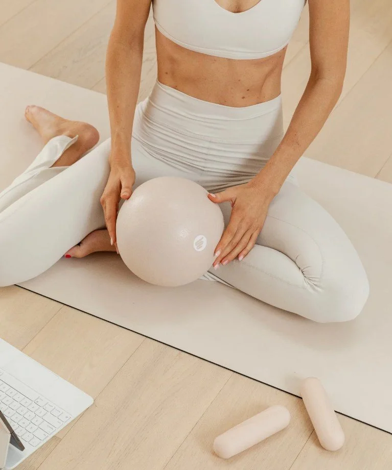 A person dressed in white workout attire sitting on a yoga mat, holding a stability ball, with exercise foam rollers on the floor nearby, in a bright workout space.