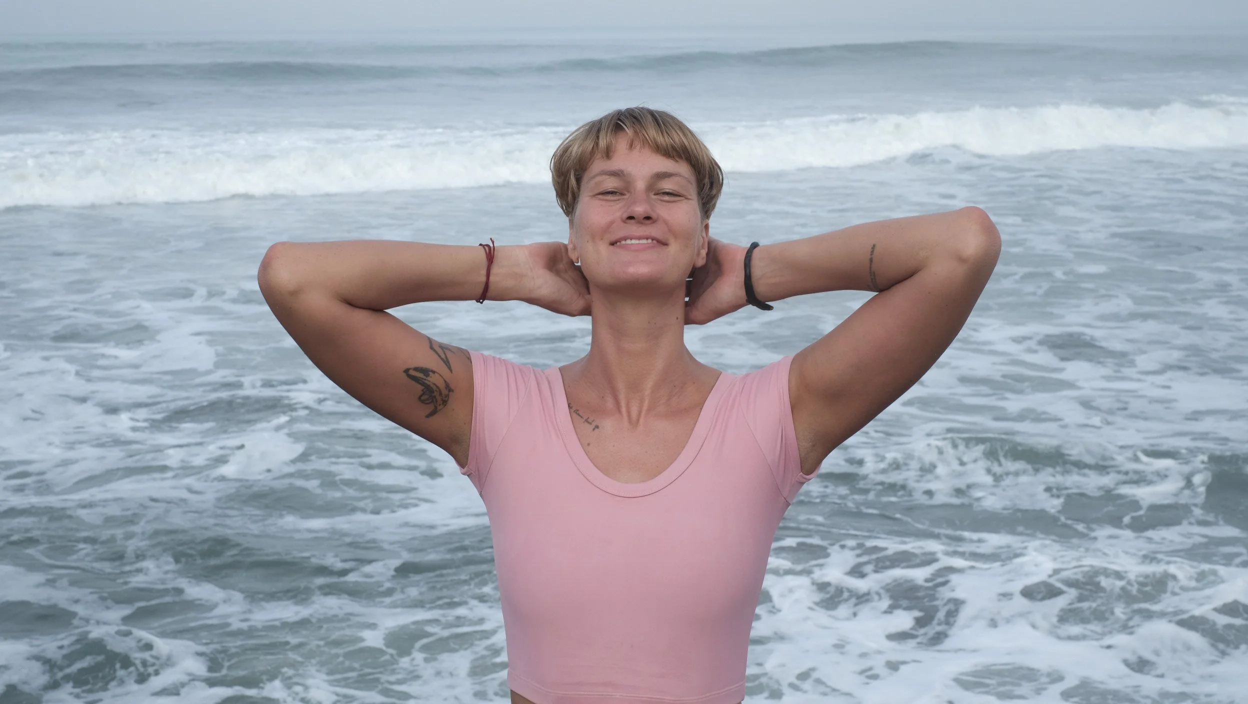 A woman with short hair smiling at the beach, standing in front of the ocean with her hands behind her head.