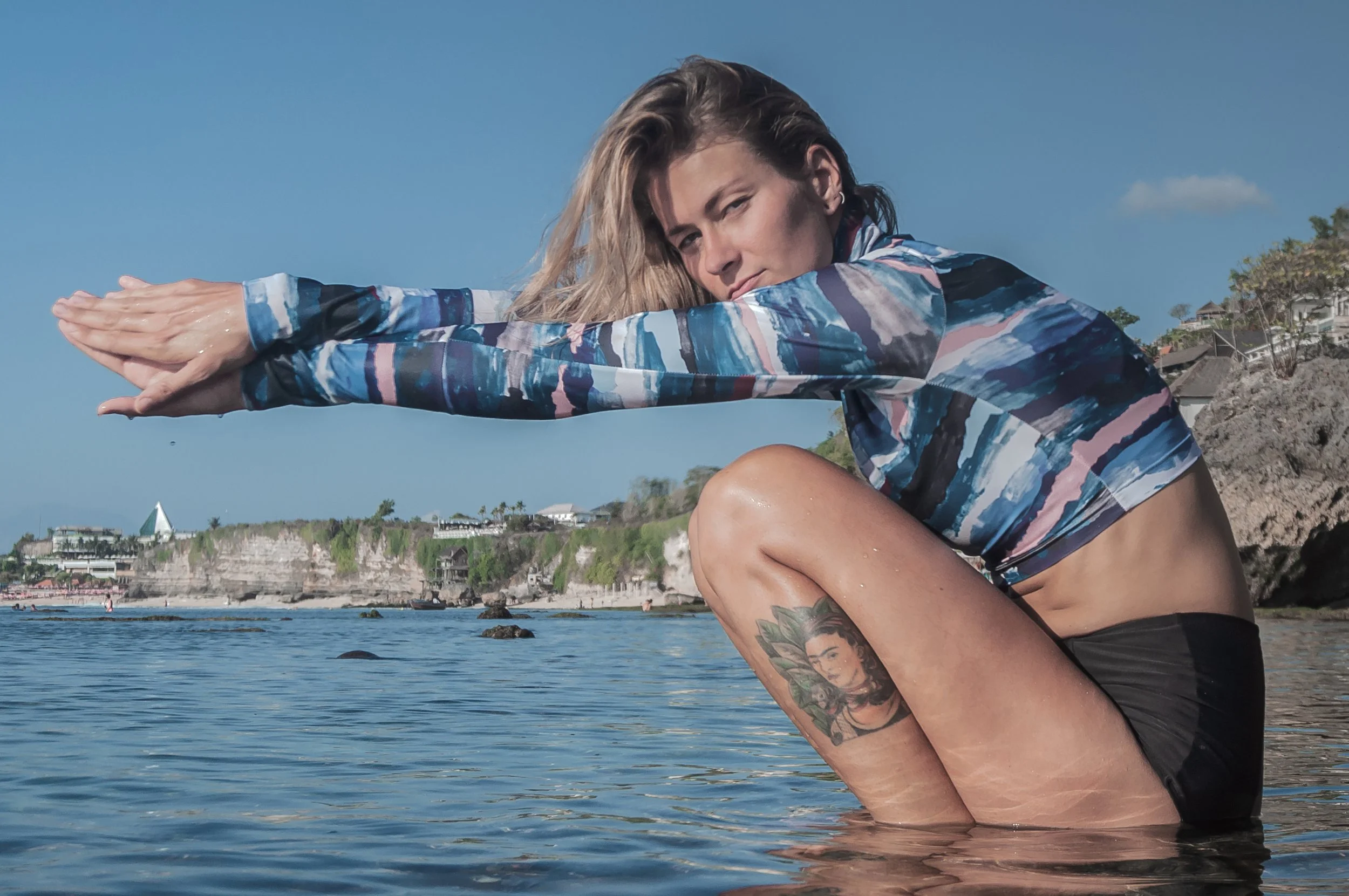 Woman in a long-sleeve athletic top and black shorts squatting in water, performing a yoga stretch on a beach with cliffs and buildings in the background.