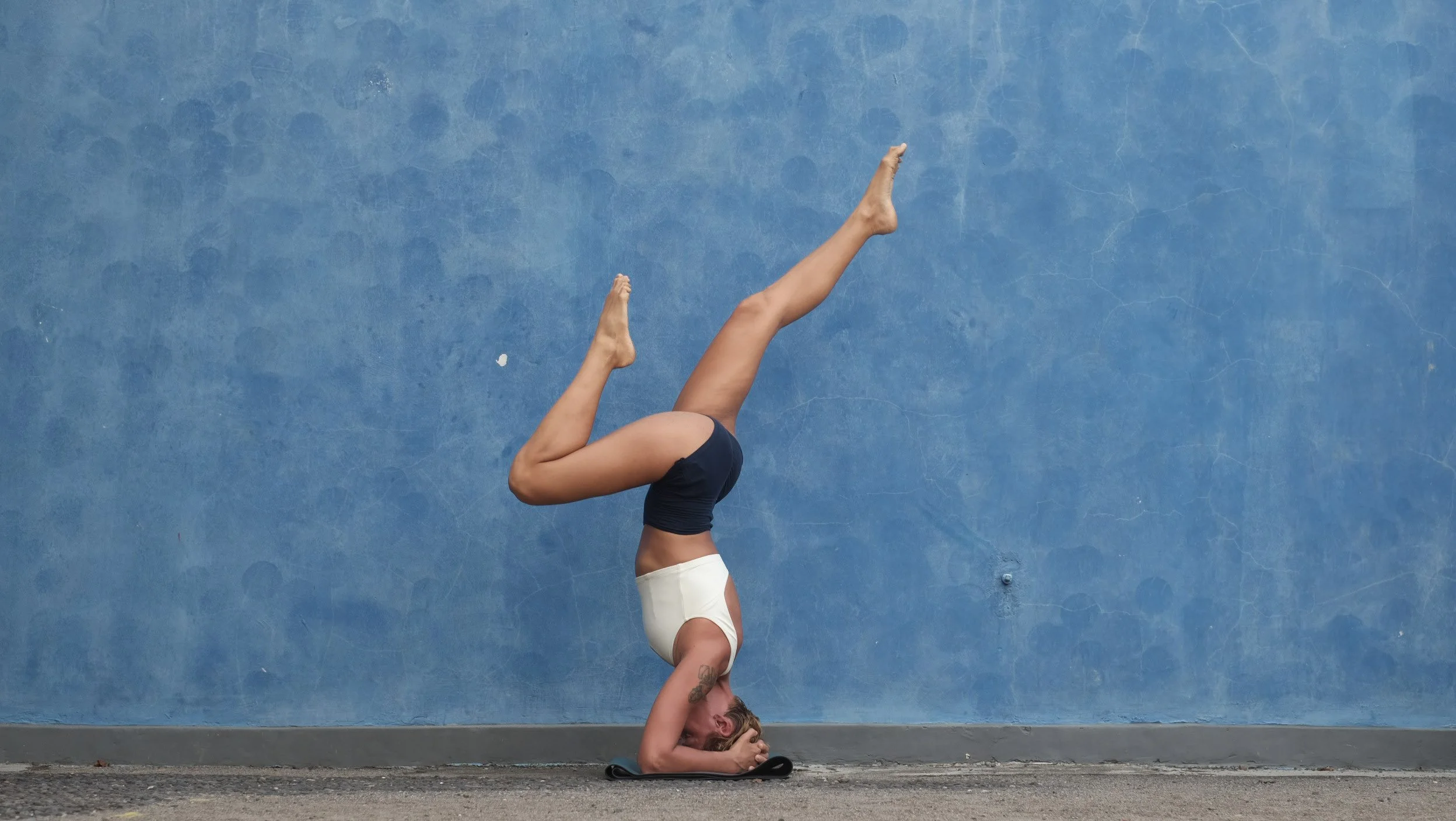 Woman practicing a headstand yoga pose against a blue wall.