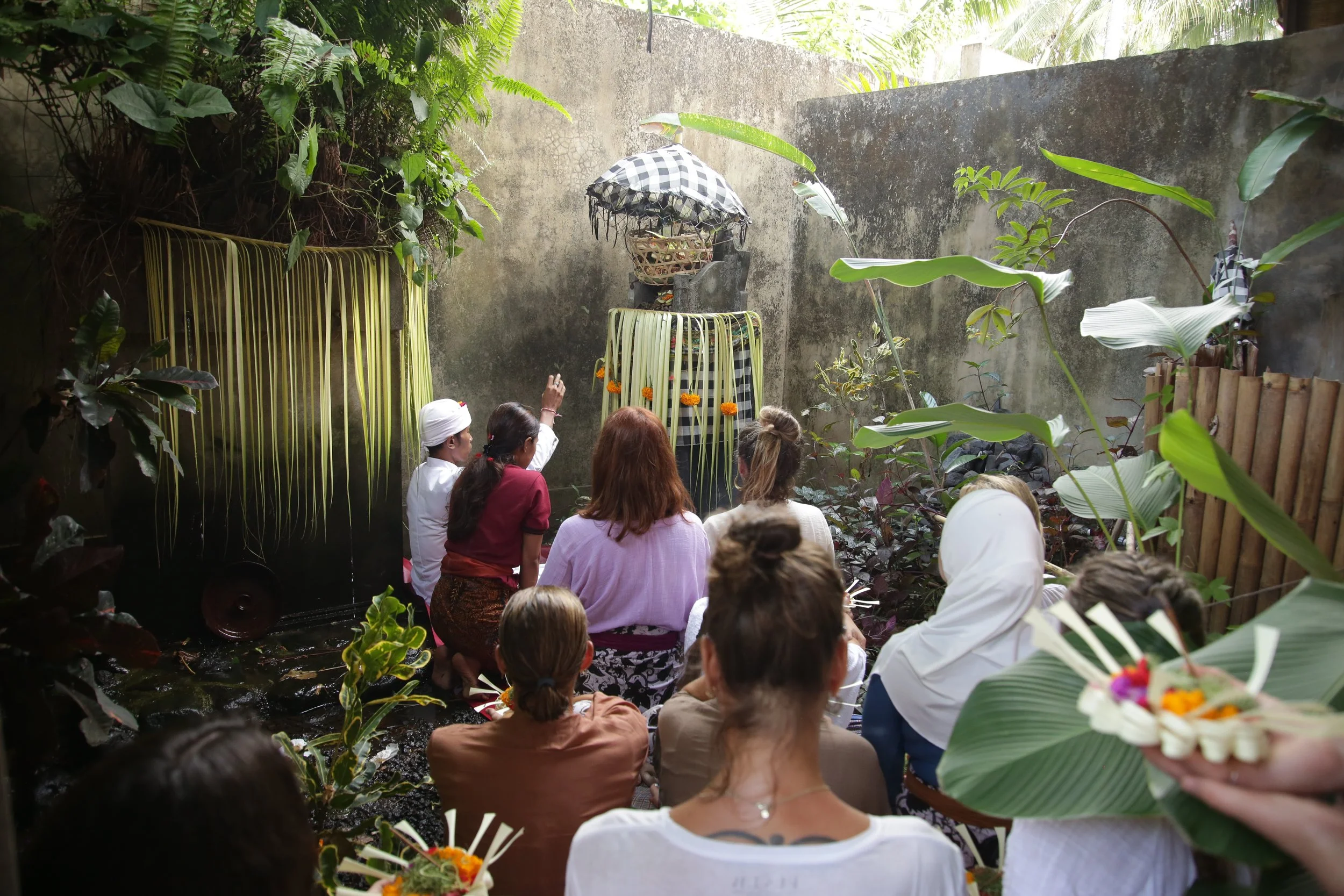 A group of people gathered in a lush, tropical garden with dense green foliage, sitting and standing around a small water feature adorned with flowers. A person dressed in traditional attire is pointing towards a structure covered with checkered fabric and decorated with marigold flowers, situated against a concrete wall. The scene appears to be a cultural or religious ceremony.