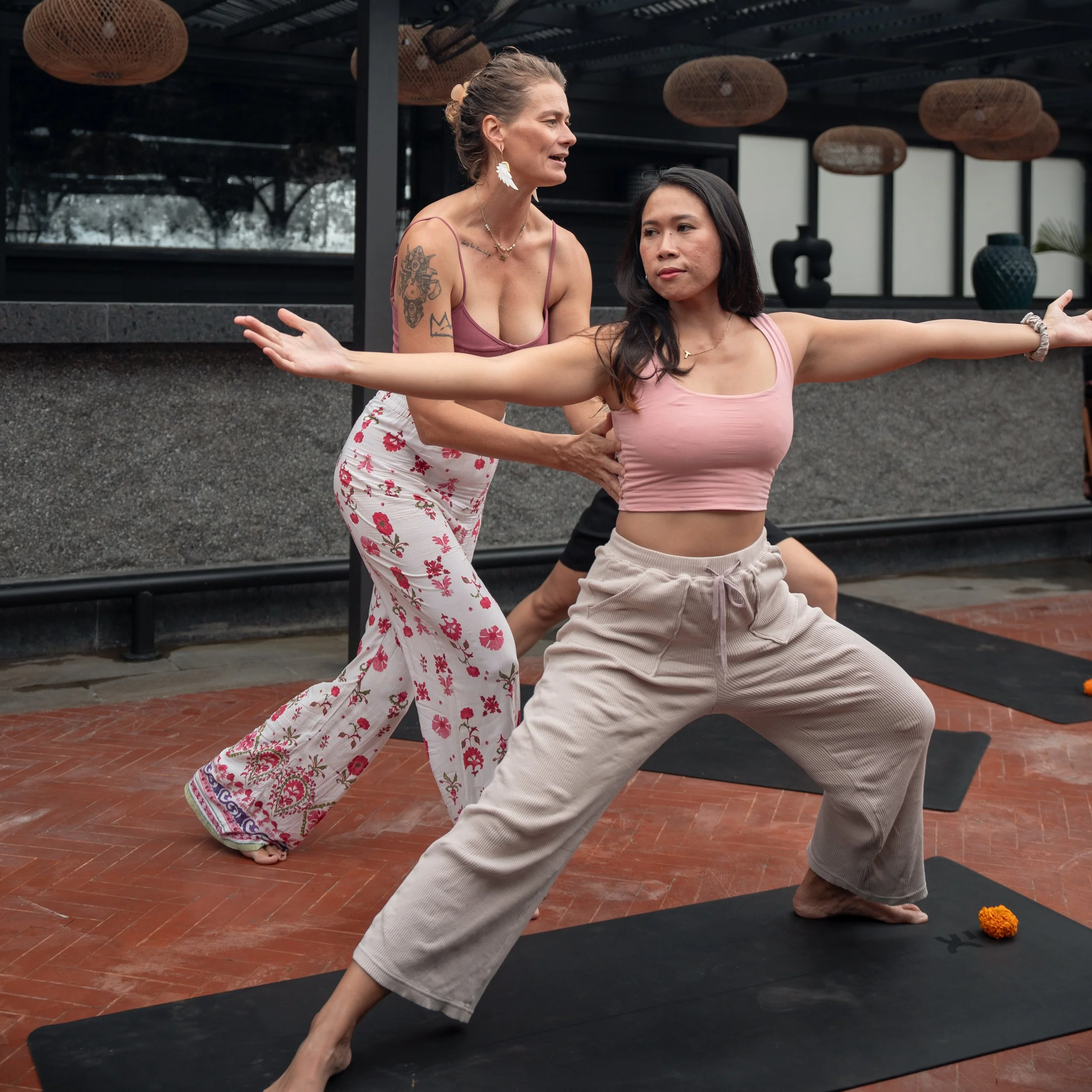 A woman with long black hair wearing a pink crop top and beige pants practicing yoga in a studio. A woman with blonde hair, tattoos, and a floral outfit provides guidance.