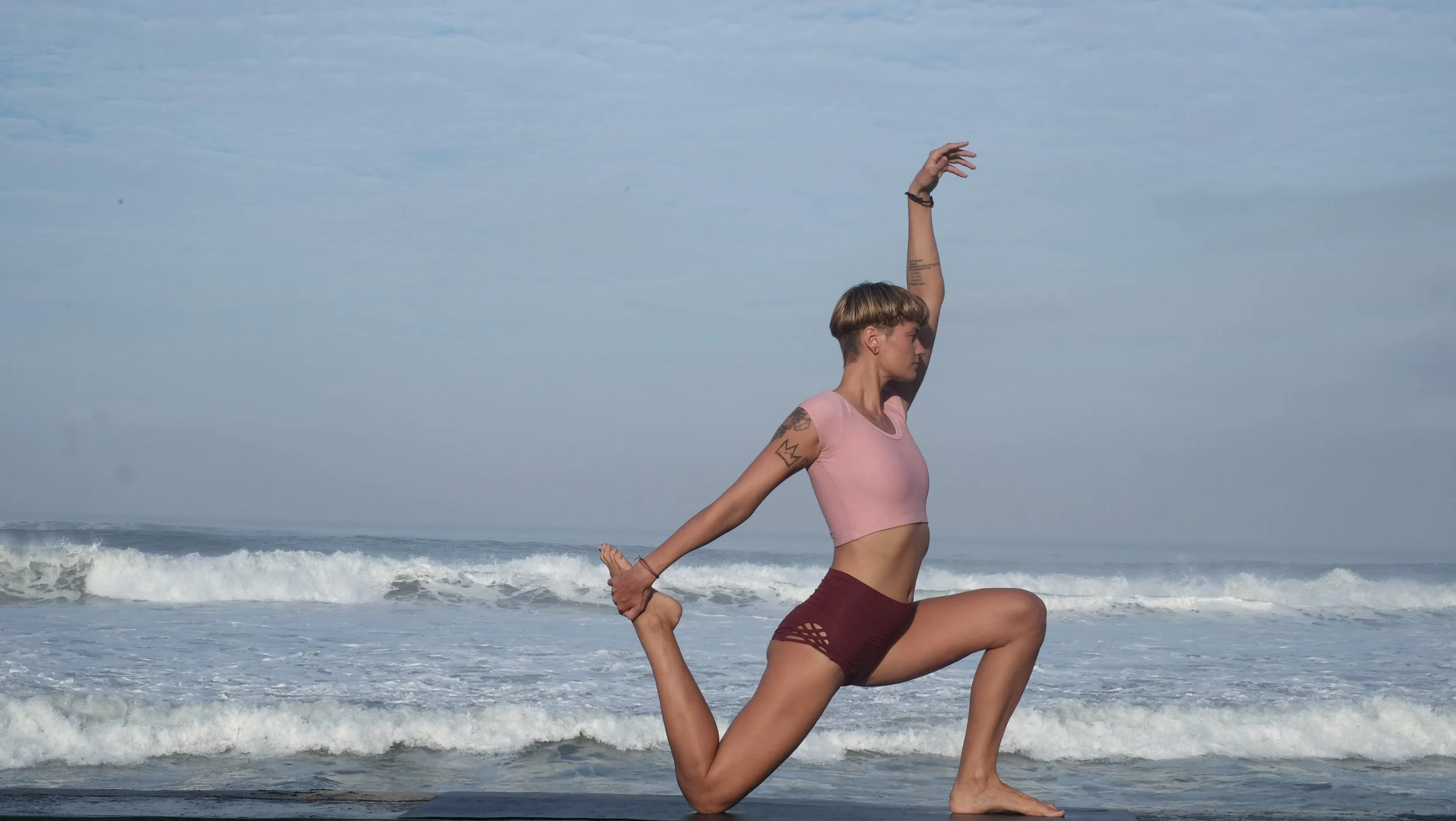 A woman practicing yoga on the beach with ocean waves in the background, performed in a lunge pose holding her back foot with her hand.