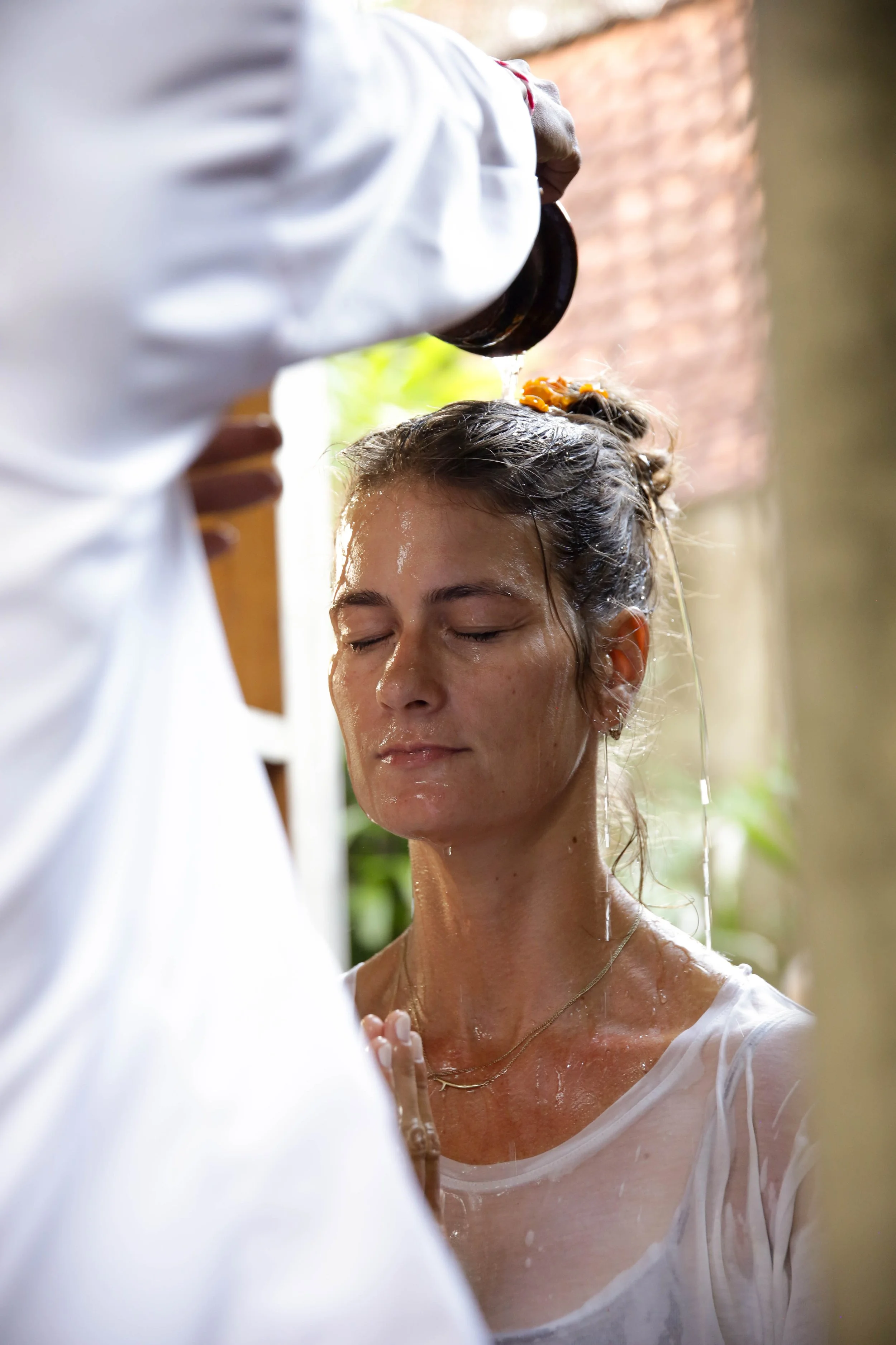 A woman with wet hair and closed eyes being baptized by a person in white robes, pouring water over her head during a baptism ceremony.