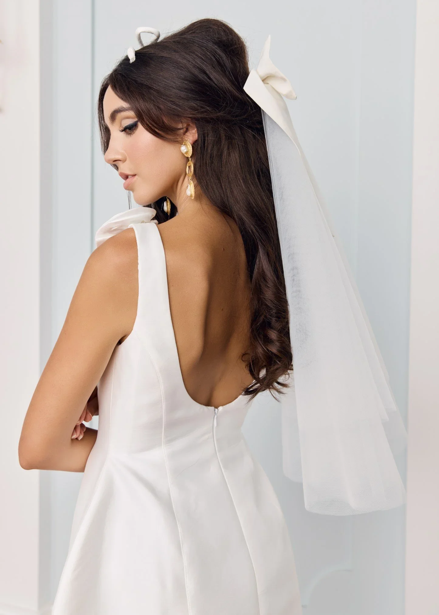 A woman wearing a white wedding dress, pearl earrings, and a long veil with a bow, standing against a neutral background.