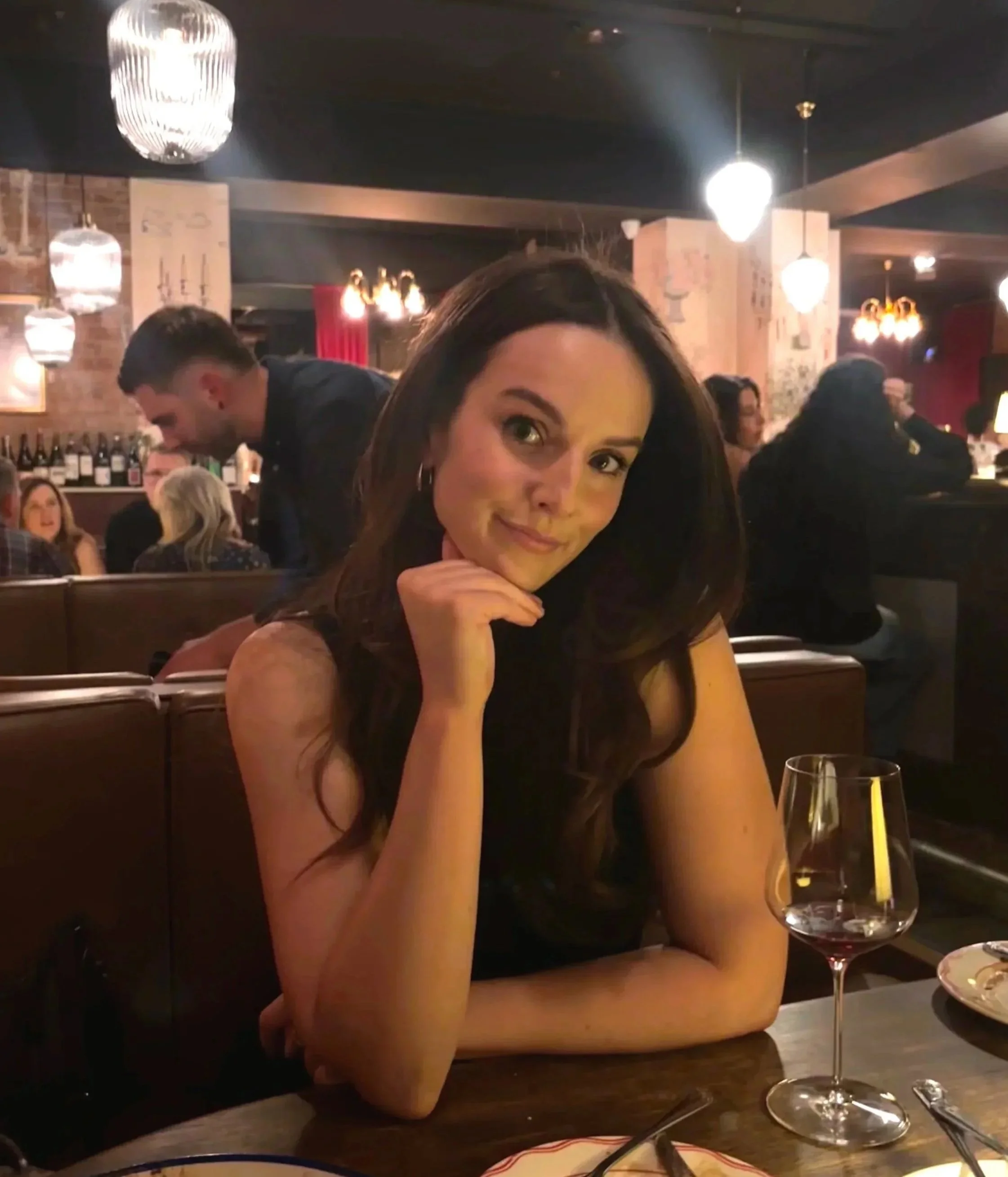 A woman sitting at a restaurant table with a glass of red wine, resting her chin on her hand and smiling at the camera.