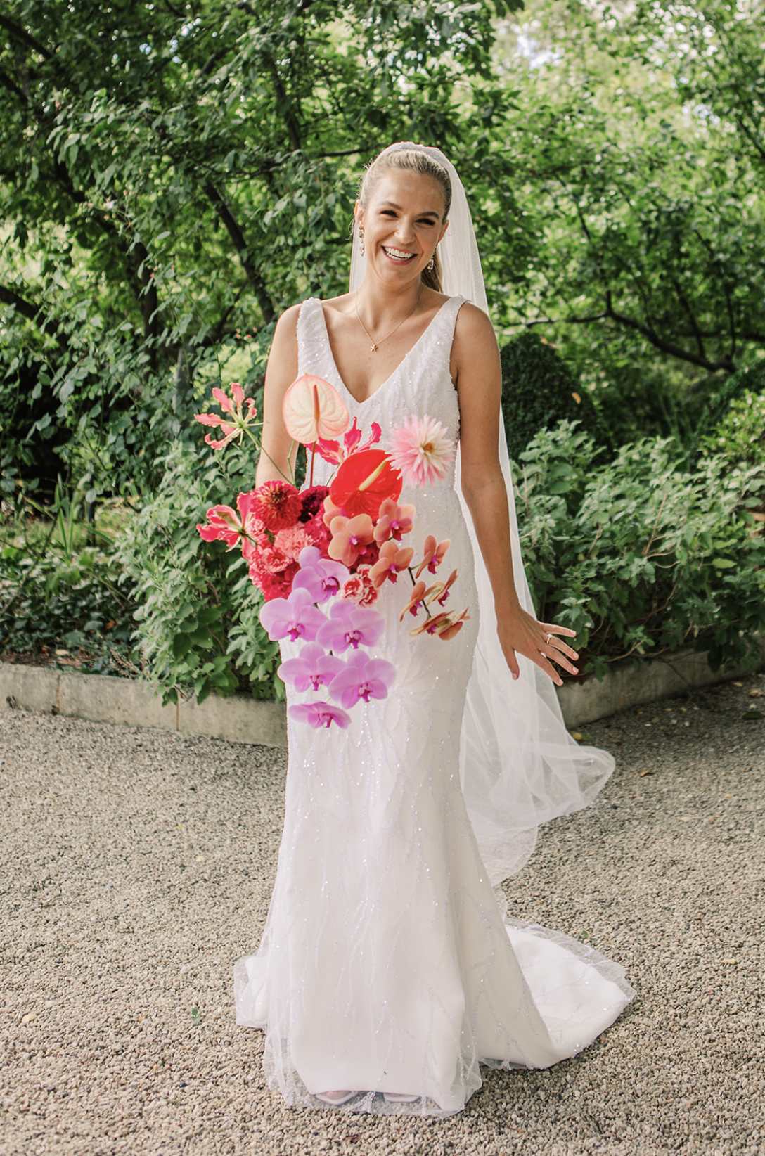 A bride in a white wedding gown smiling and holding a large bouquet of pink and red flowers. She is standing outdoors on a gravel path with green foliage in the background.