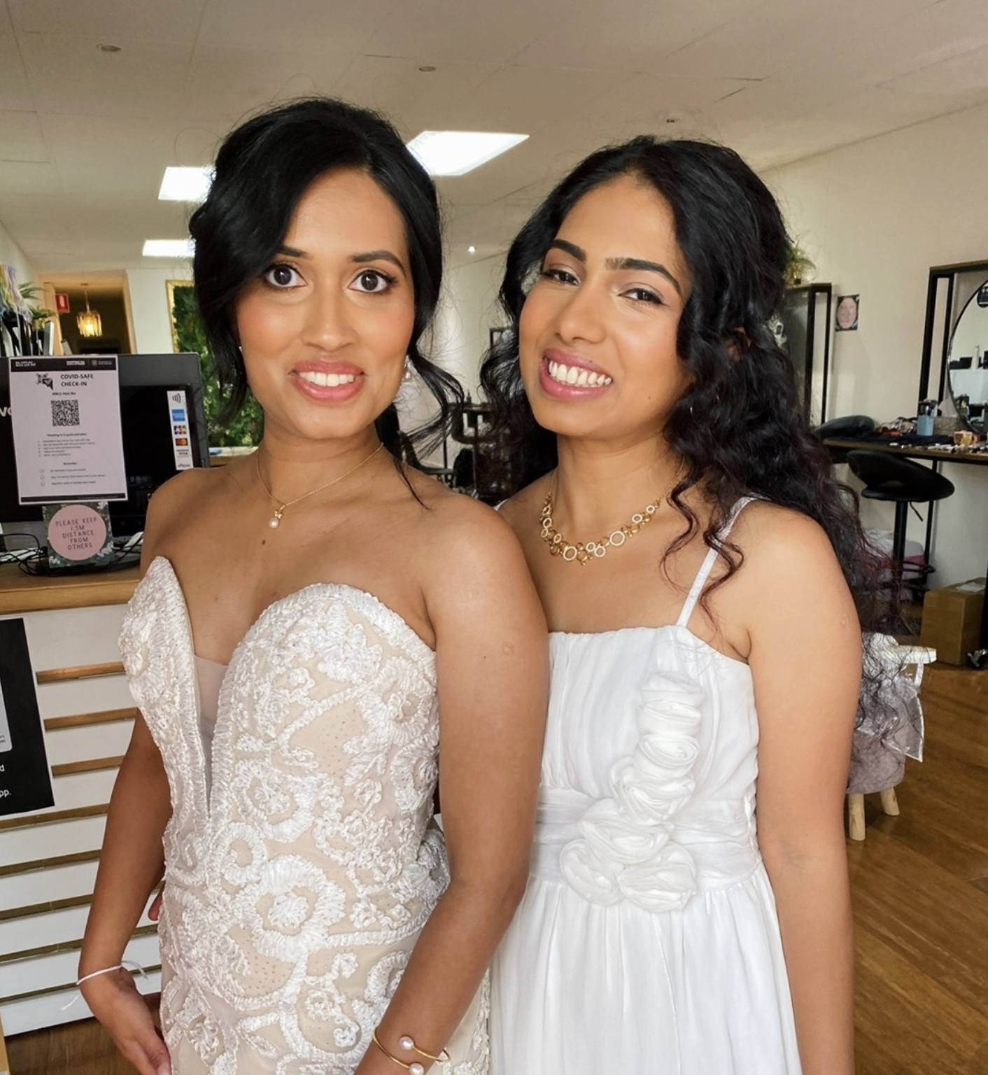 Two women wearing white dresses, smiling, standing close together indoors.
