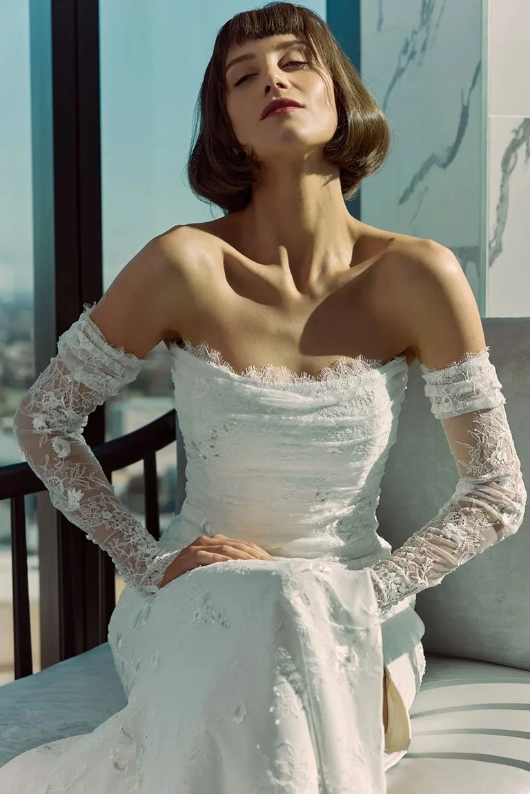 A woman in a white lace off-shoulder dress sitting on a sofa near a window.