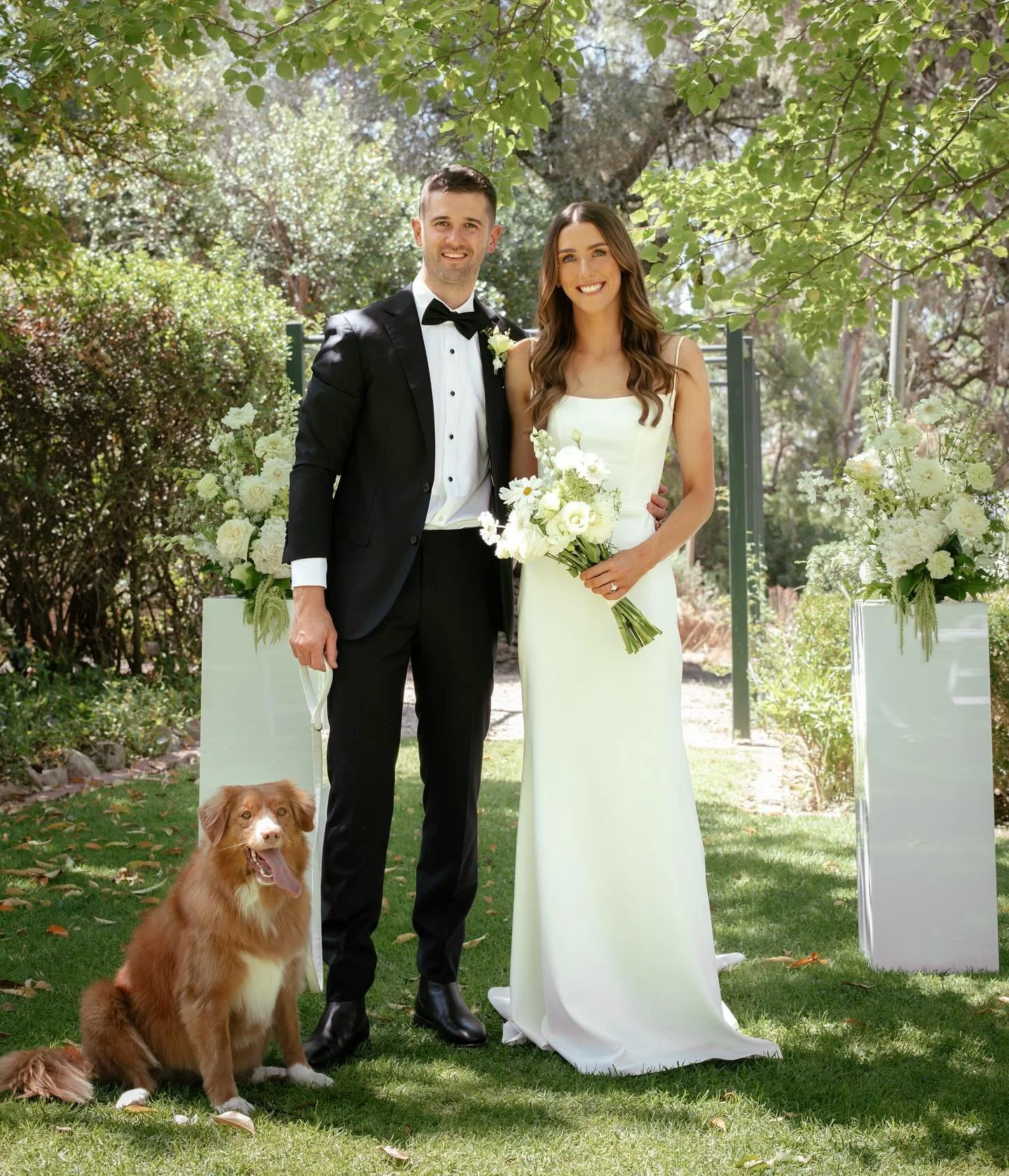 A bride and groom standing outdoors under a tree, smiling, with a brown dog sitting on the grass beside them, surrounded by white floral arrangements.