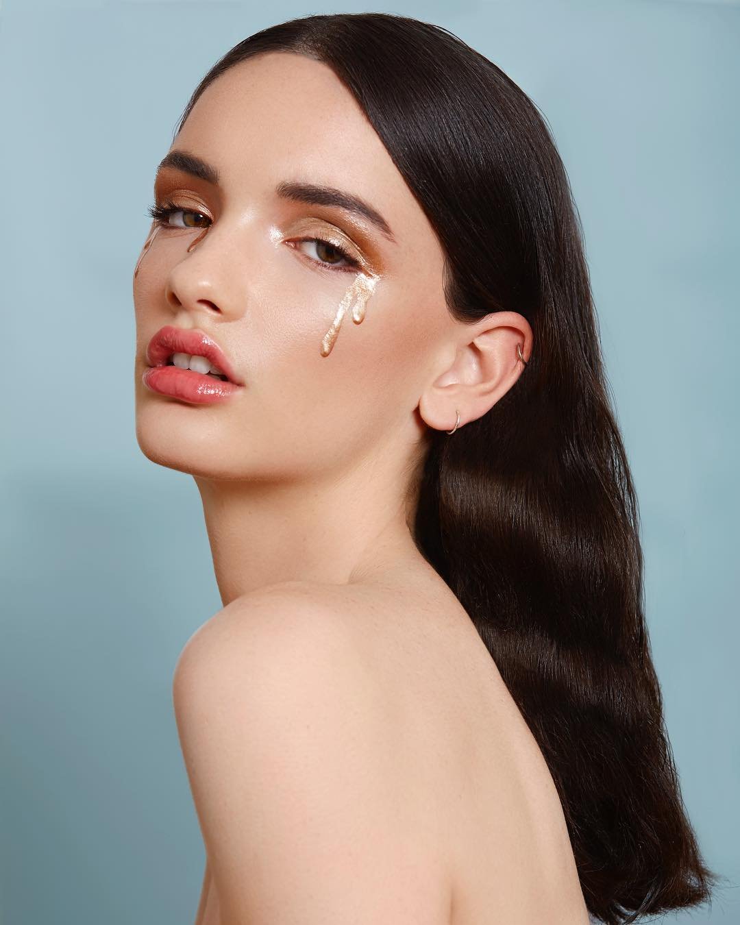 A woman with long dark hair and fair skin, wearing makeup and a subtle expression, with a gold tear-shaped embellishment under her eye against a light blue background.