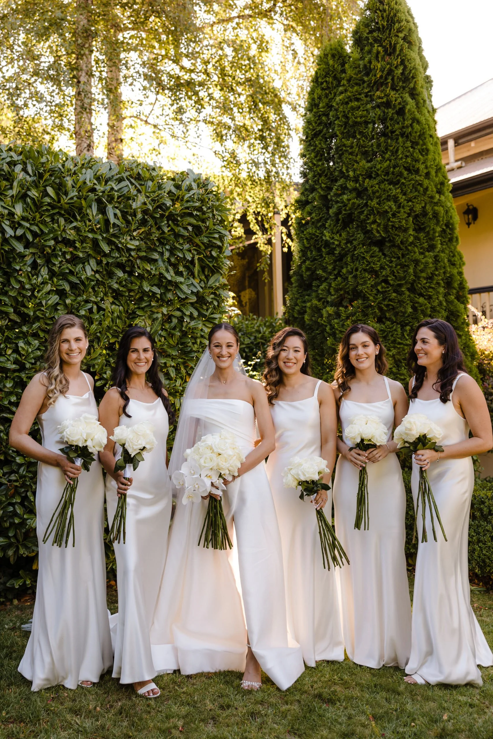 A group of six women dressed in white bridesmaid dresses, standing outdoors in a garden, holding white bouquets, celebrating a wedding.