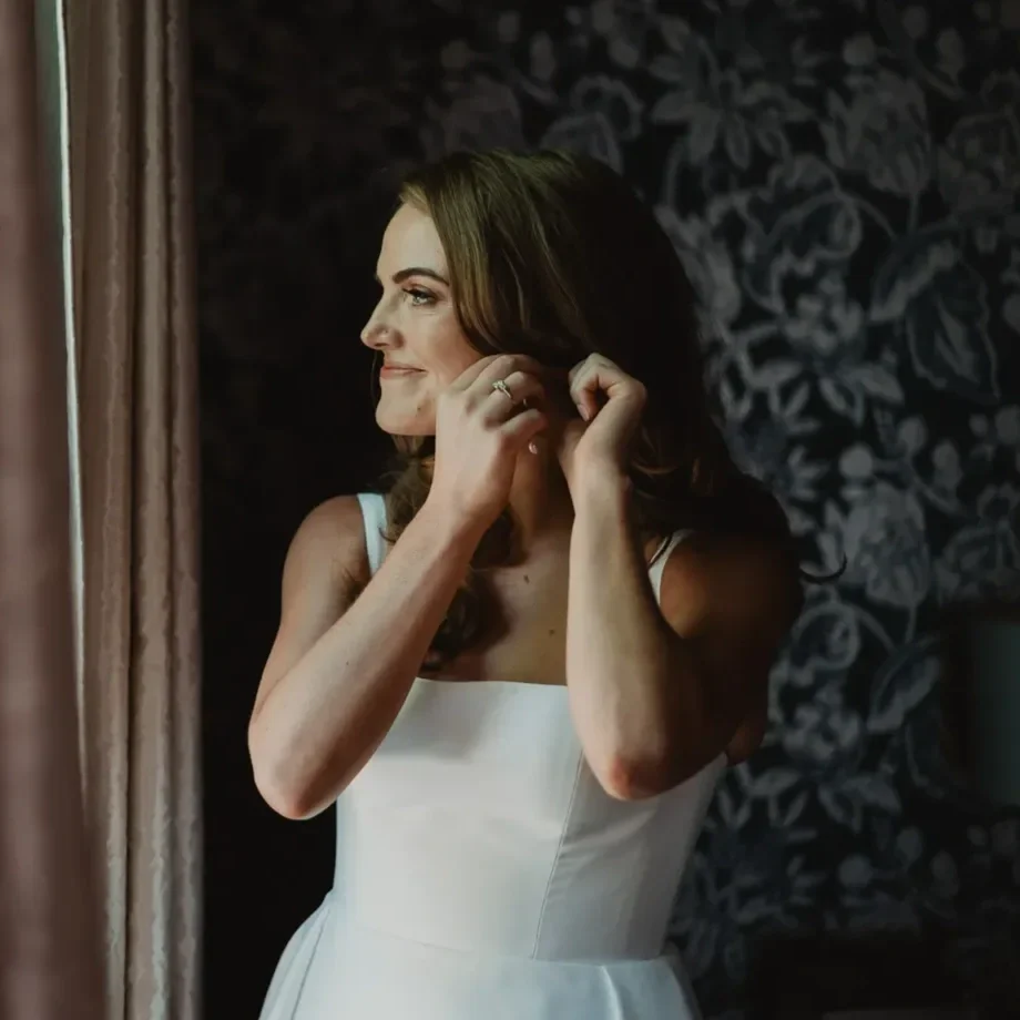 A woman in a white dress is fixing her earring while smiling, standing near a window with beige curtains. There is dark floral wallpaper behind her.