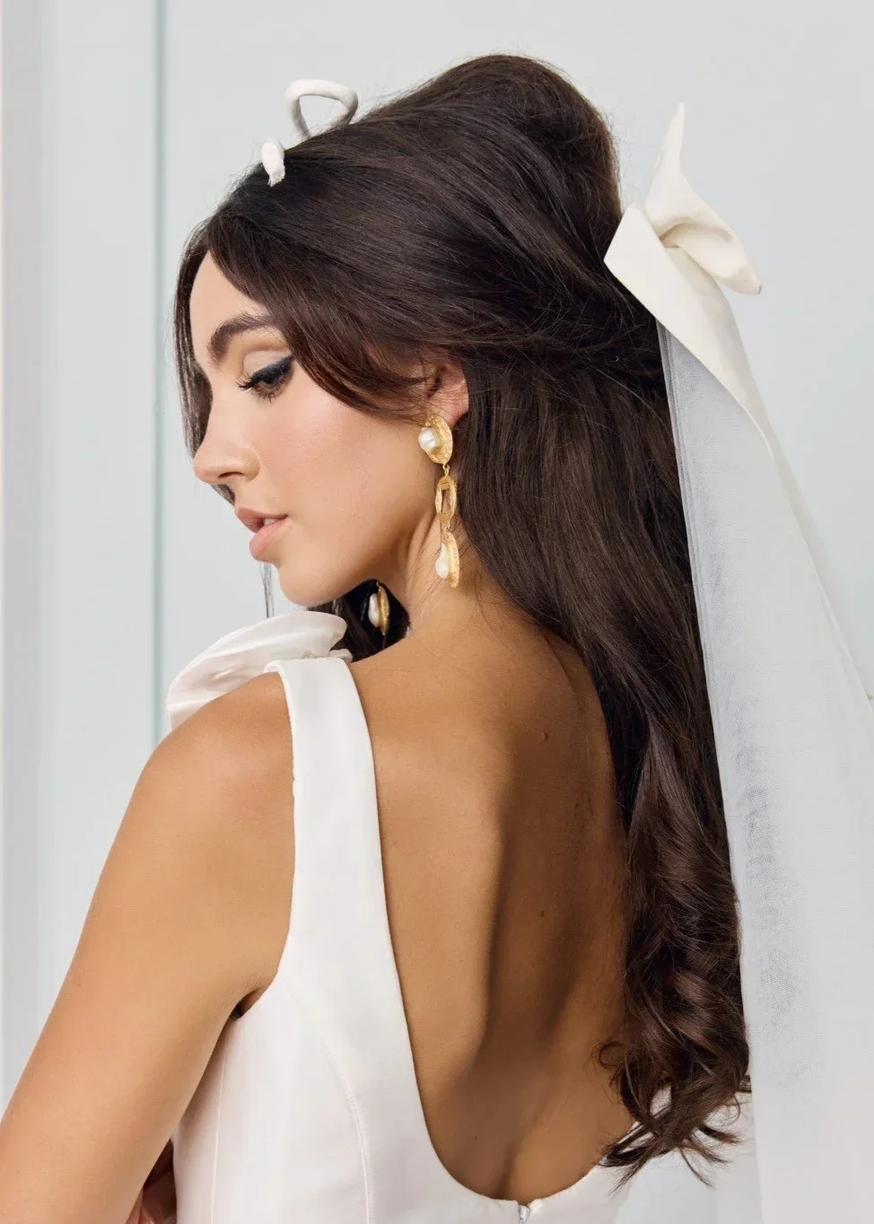A woman with dark brown hair styled in loose waves, wearing elegant pearl and gold earrings, a white dress with a low back, and a white bow in her hair, looking downward.