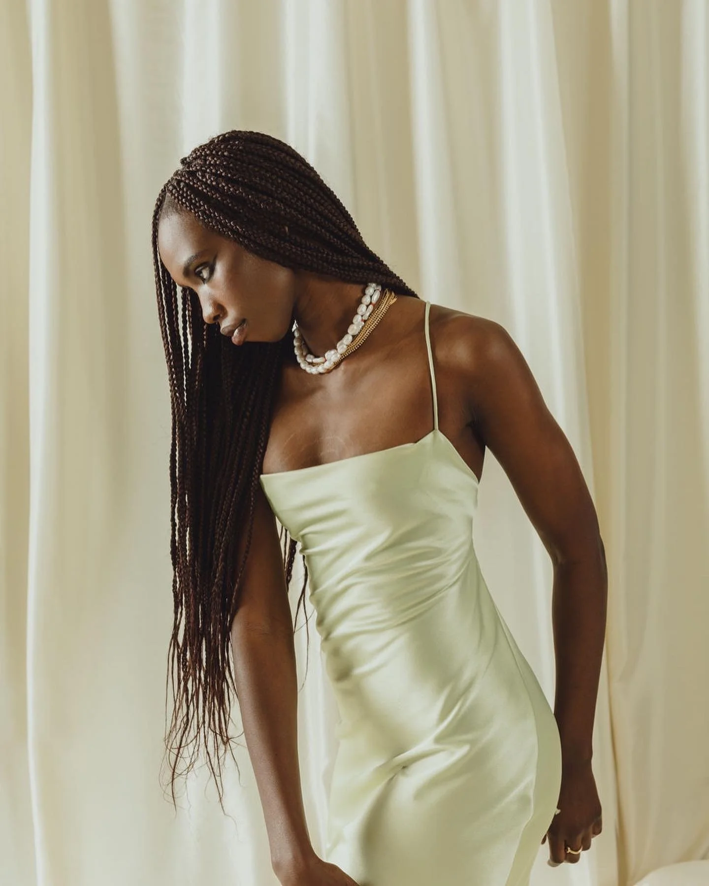A woman with braided hair wearing a silky, light-colored dress and pearl jewelry standing in front of cream-colored curtains.