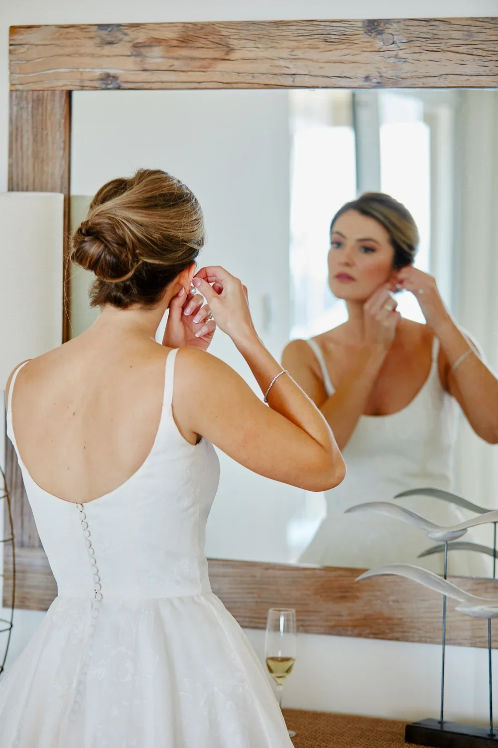 A bride wearing a white wedding dress is seen from behind, adjusting her earring while looking at her reflection in a large mirror.