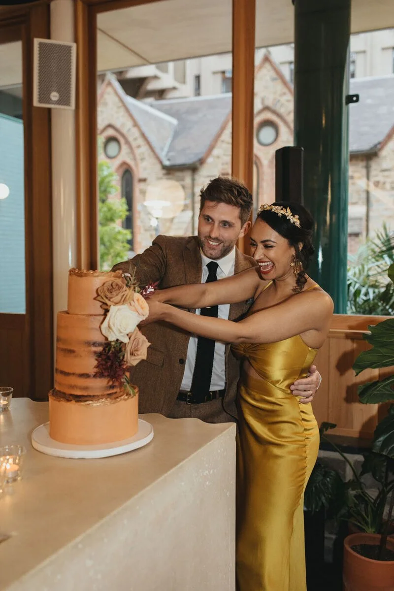 A couple cutting a wedding cake together at their celebration, with the bride wearing a gold dress and tiara, and the groom in a brown suit, in a warmly decorated indoor setting.