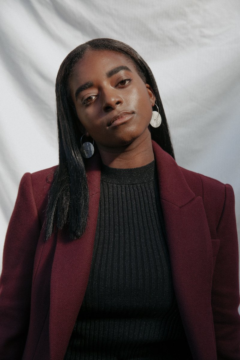 A young woman with dark hair and skin, wearing a burgundy blazer and a black ribbed top, with earrings, posing in front of a white backdrop.
