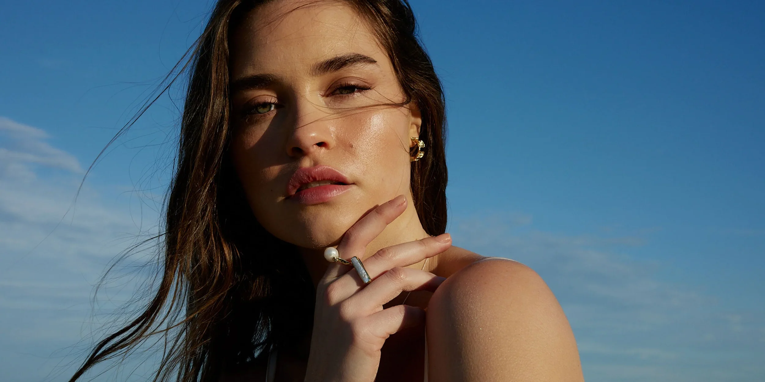 Close-up of a woman with brown hair, wearing gold earrings and rings, posing outdoors against a blue sky background.