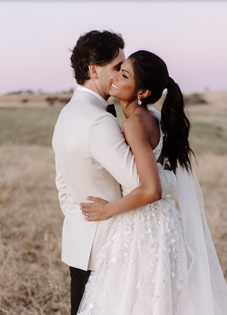 A bride and groom embrace outdoors during sunset, smiling with eyes closed, in a field with distant hills.