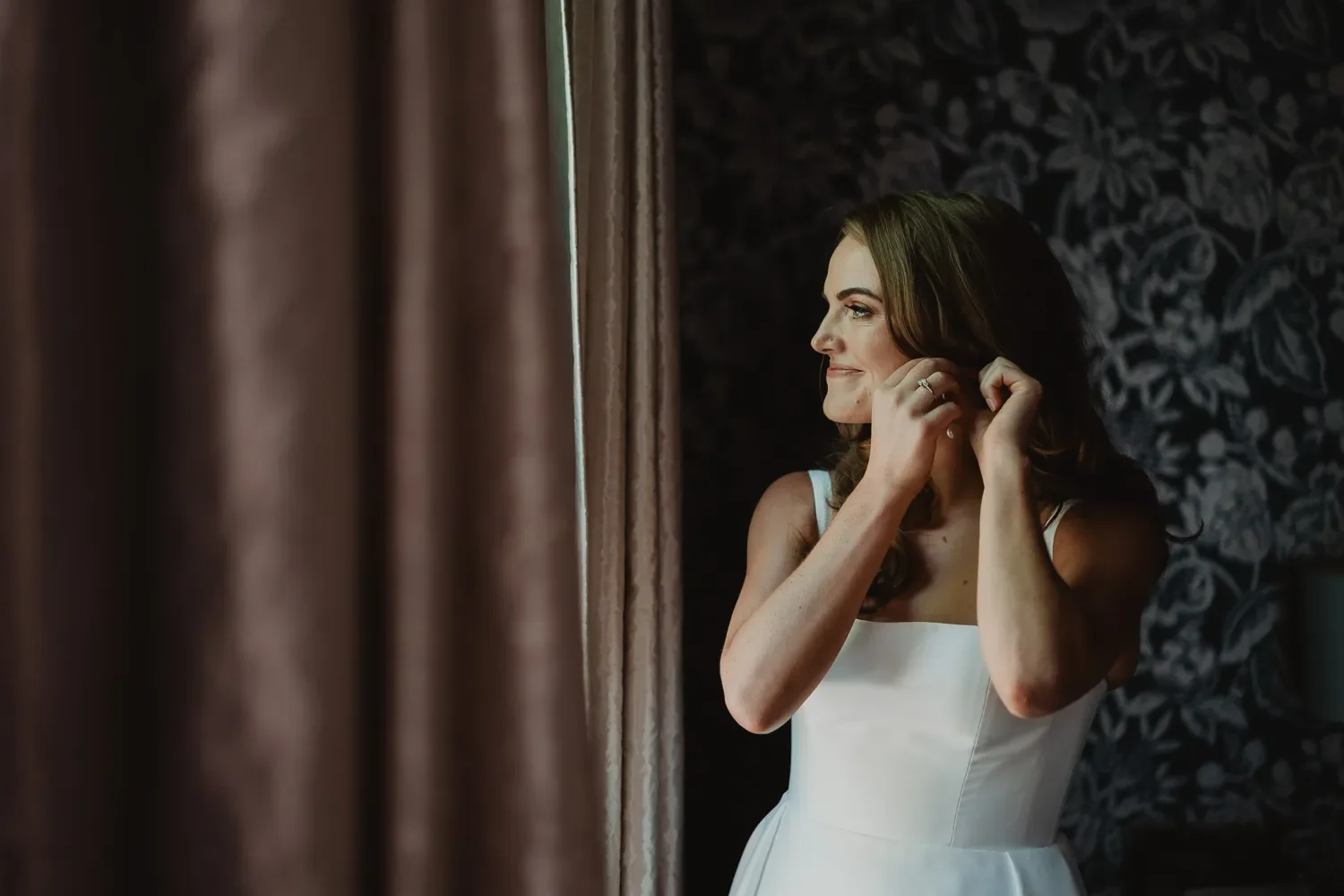 A woman in a white dress is putting on earrings next to a window with curtains, with a dark patterned wall in the background.