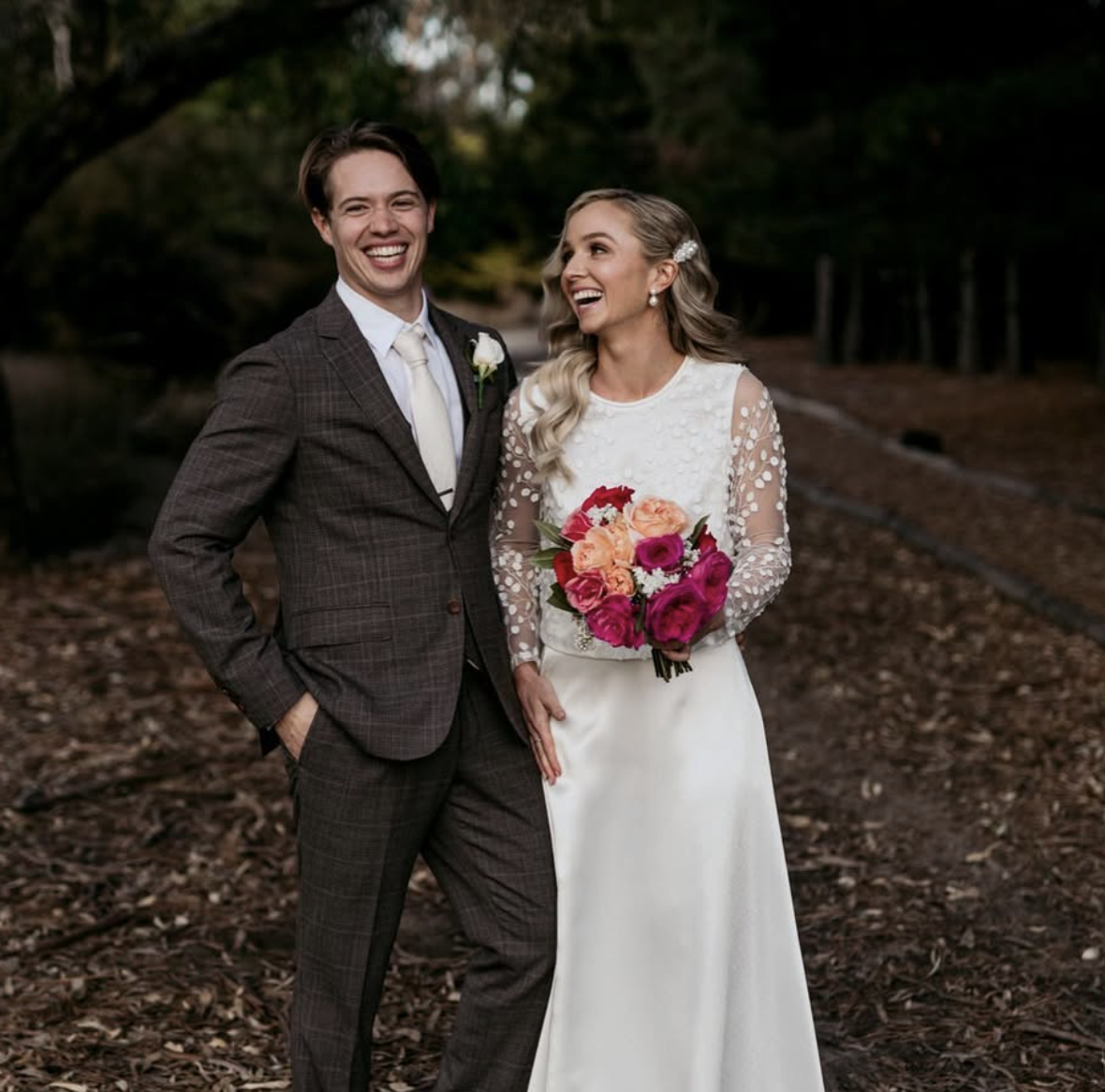 A happy bride and groom standing outdoors on a wooded path with leaf-covered ground, smiling and laughing, with trees in the background. The bride is holding a bouquet of colorful flowers and wearing a white wedding dress with lace sleeves. The groom