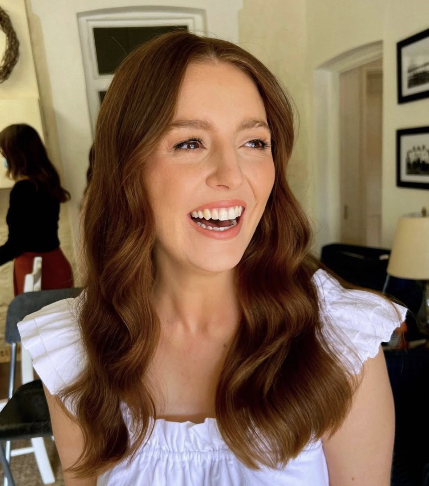 A woman with long, wavy red hair, smiling with her mouth open, wearing a white top with ruffled sleeves.