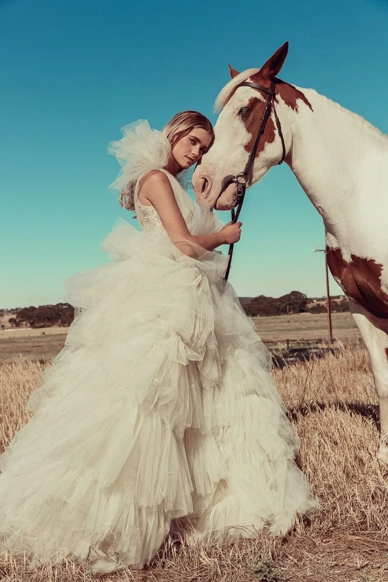 A woman in a fancy white dress with tulle layers standing in a field, holding the reins of a white horse with brown patches, under a clear blue sky.
