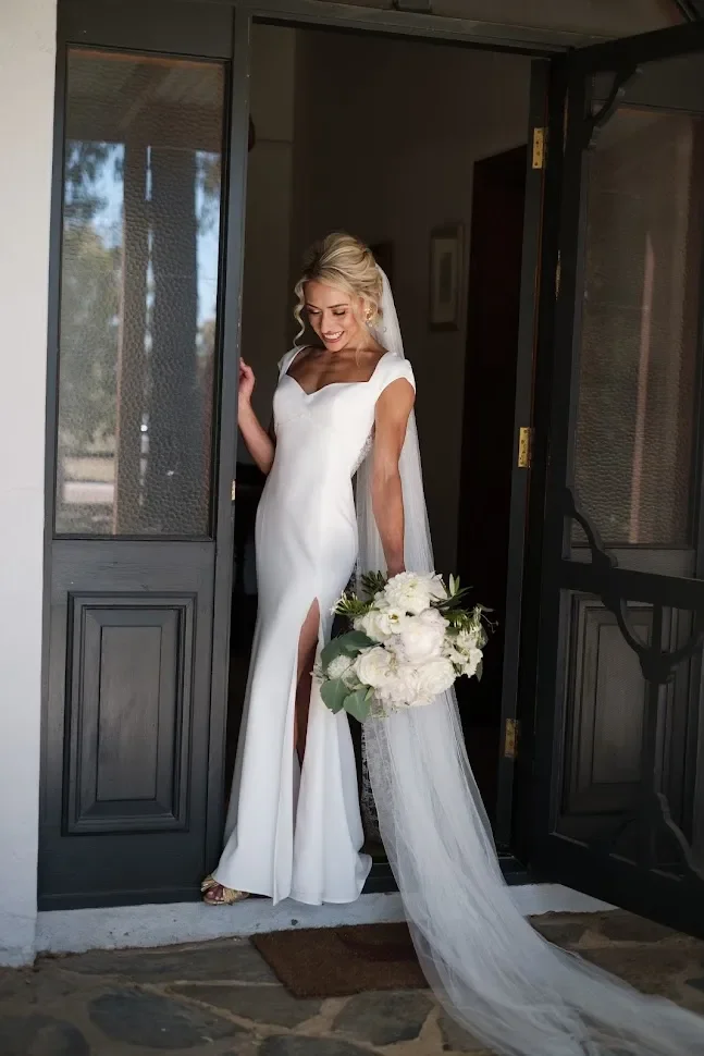 A bride in a white wedding dress holding a bouquet of white flowers while standing in a doorway, smiling and looking down.