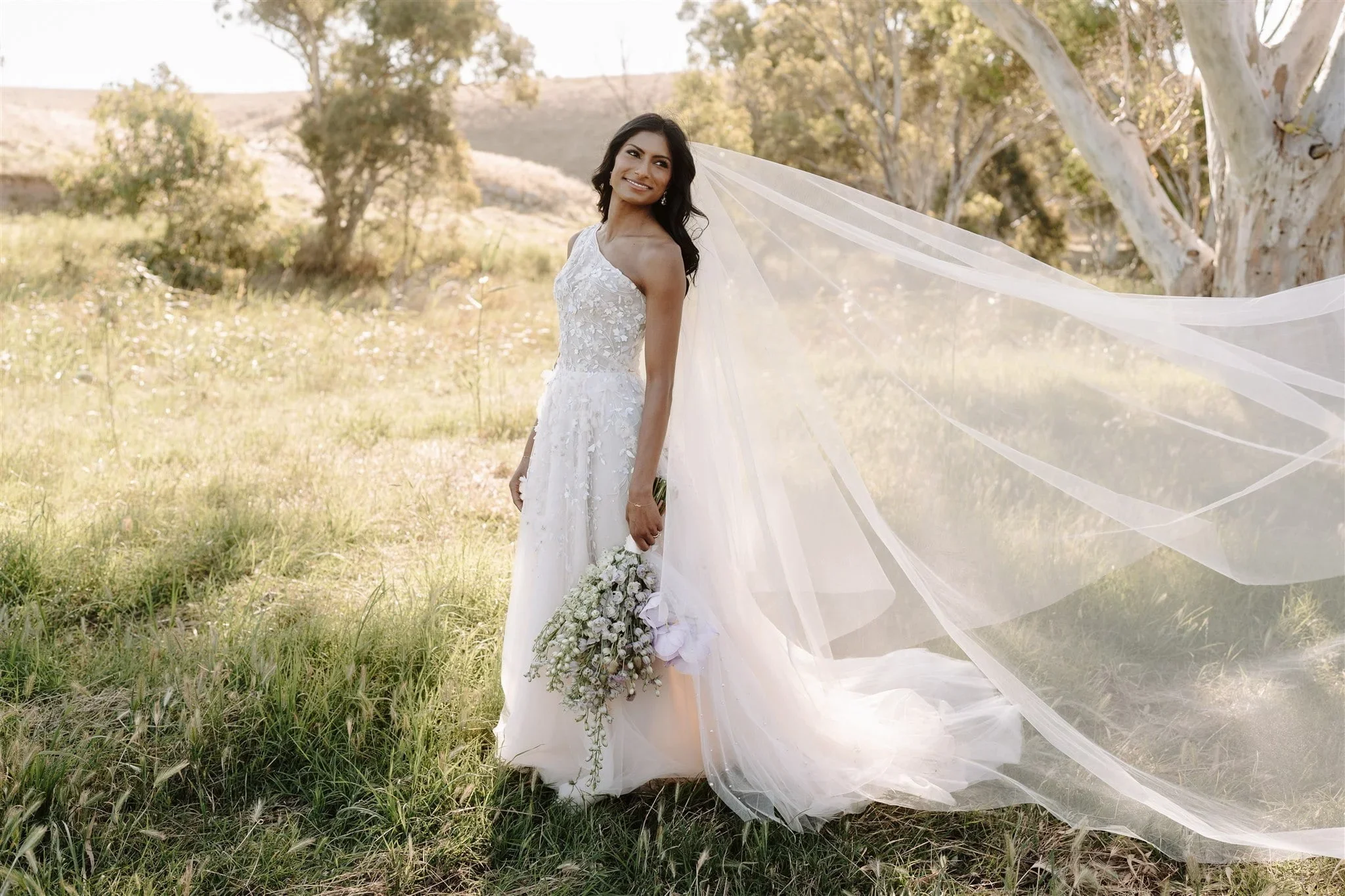 A bride in a white wedding dress standing outdoors in a grassy area with trees, holding a bouquet of white flowers, smiling, with her veil flowing behind her.