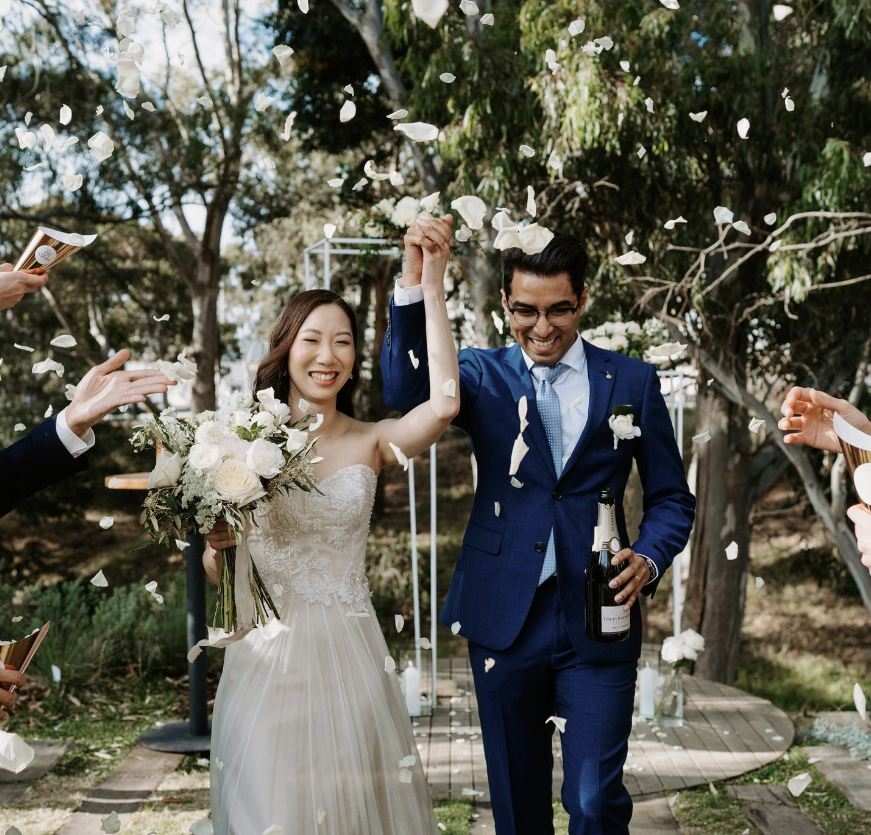 Happy bride and groom celebrating outdoors after wedding, holding hands up amidst falling flower petals, with wedding guests around, and trees in the background.