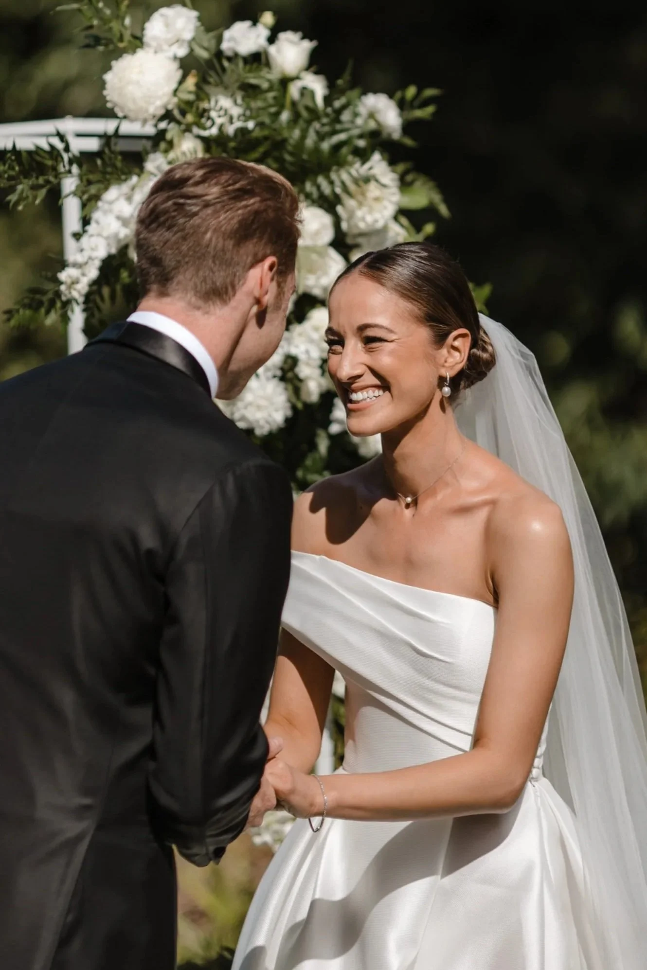 A bride smiling happily at her groom during their wedding ceremony, with white flowers in the background.