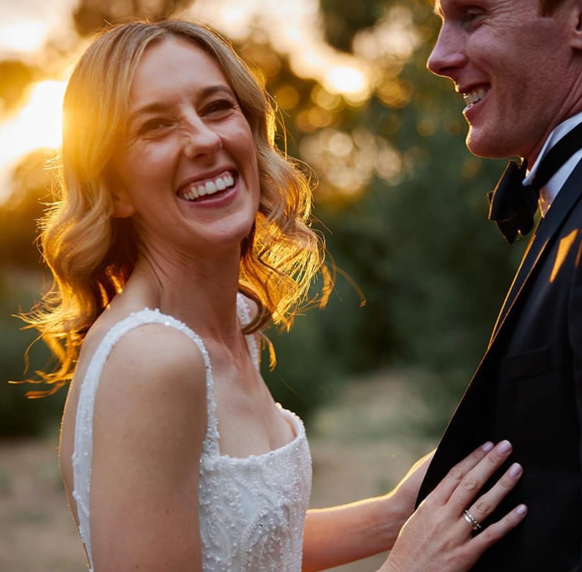A smiling bride in a white dress and a groom in a tuxedo sharing a joyful moment outdoors during sunset.