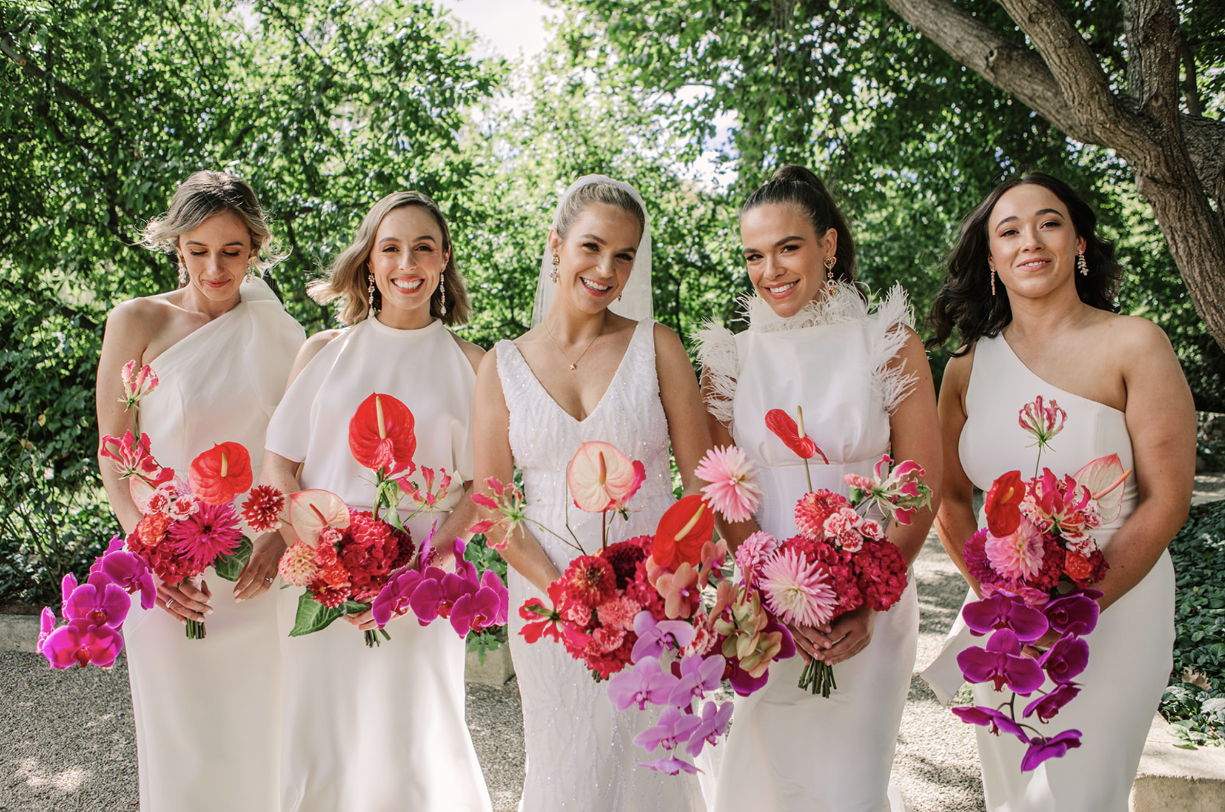 Bride and five bridesmaids standing outdoors under trees, all holding bouquets of pink and red flowers.