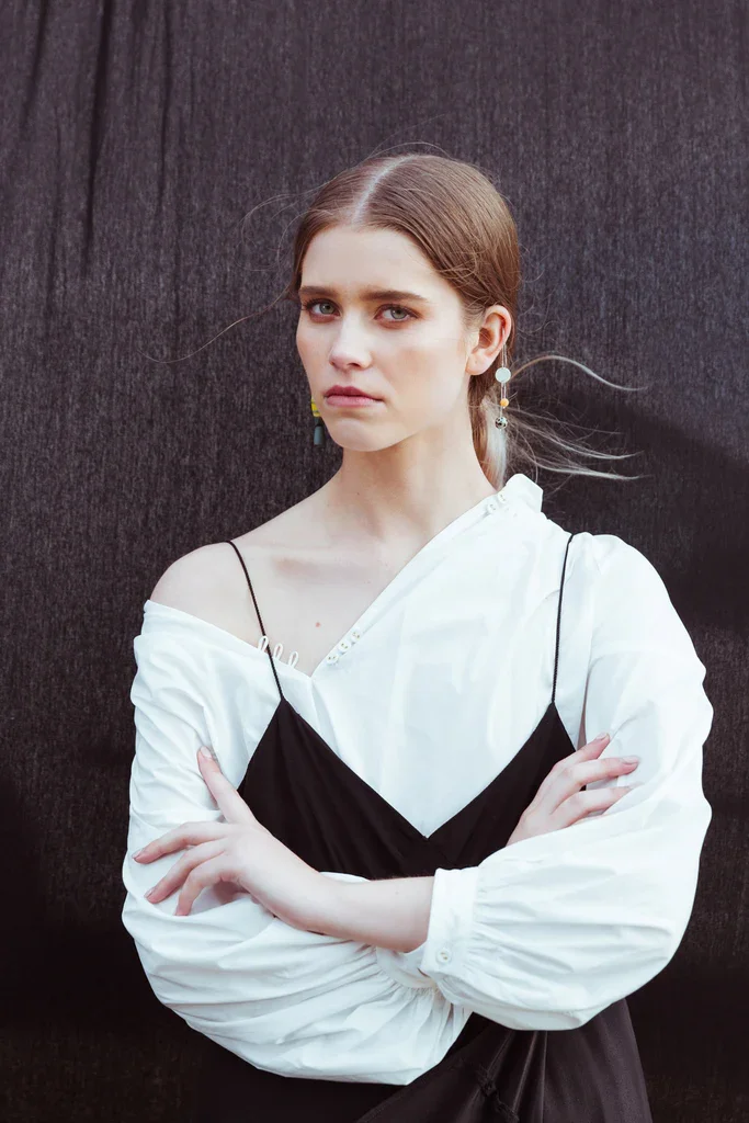 A young woman with long, straight, reddish-brown hair posing against a dark background. She is wearing a white and black layered top with off-the-shoulder and spaghetti straps, with her arms crossed.