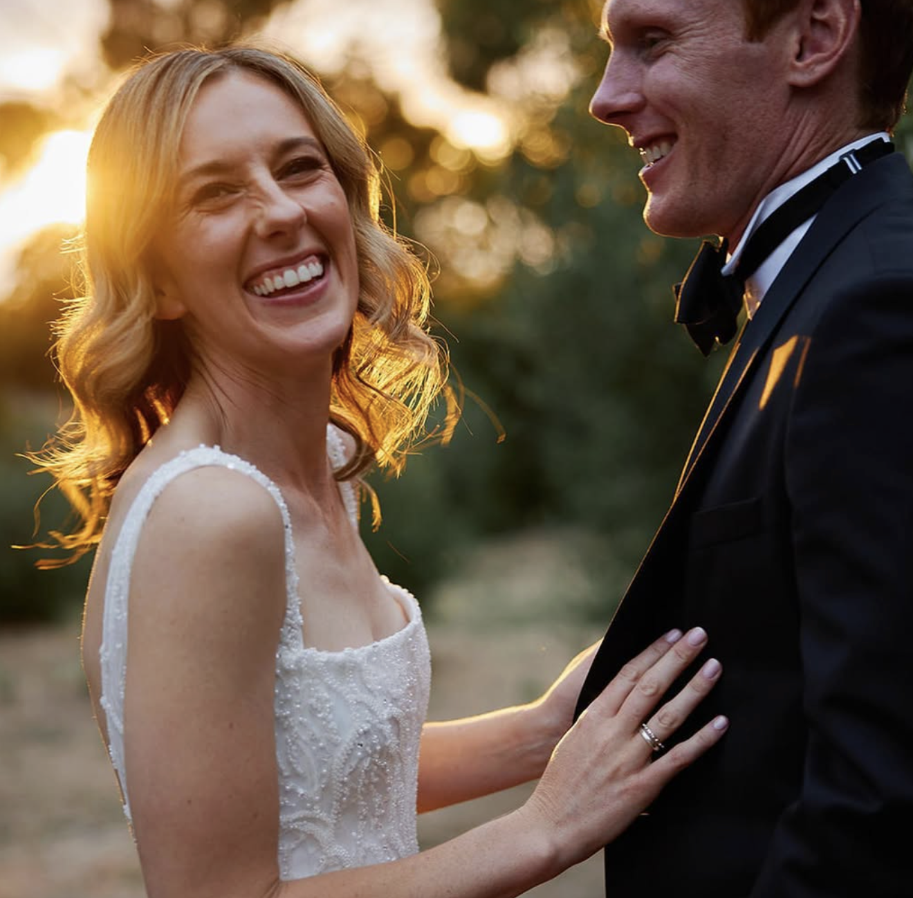 A woman in a white wedding dress smiling and holding hands with a man in a black tuxedo at sunset outdoors.