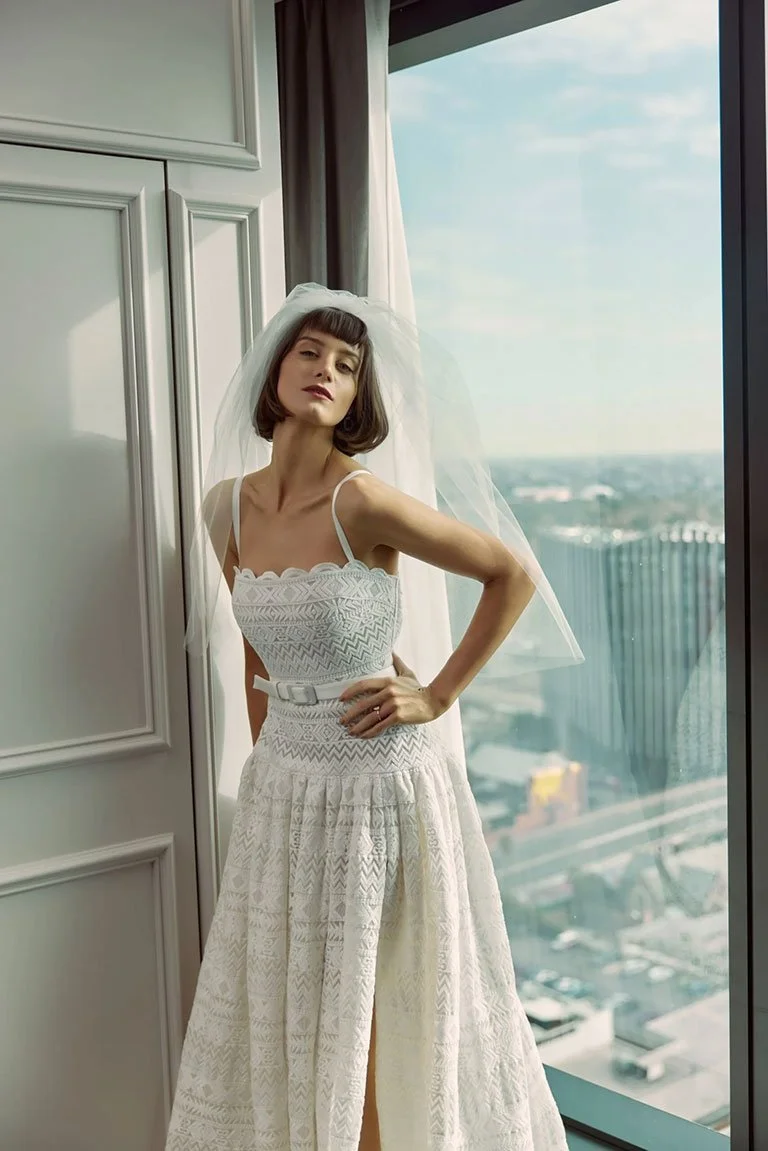 A woman in a white dress with a veil standing in front of a large window with a city view.