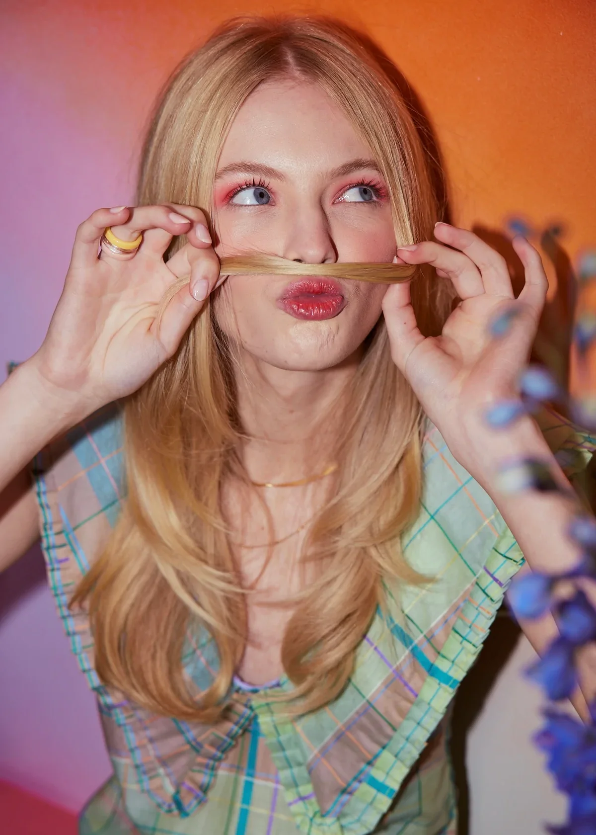 A young woman with long blonde hair making a funny face, holding a strand of her hair under her nose like a mustache, with pink and orange background.