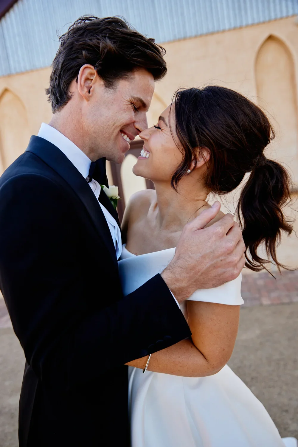A bride and groom are smiling and touching foreheads, embracing outdoors, with the bride in a white wedding dress and the groom in a black tuxedo.