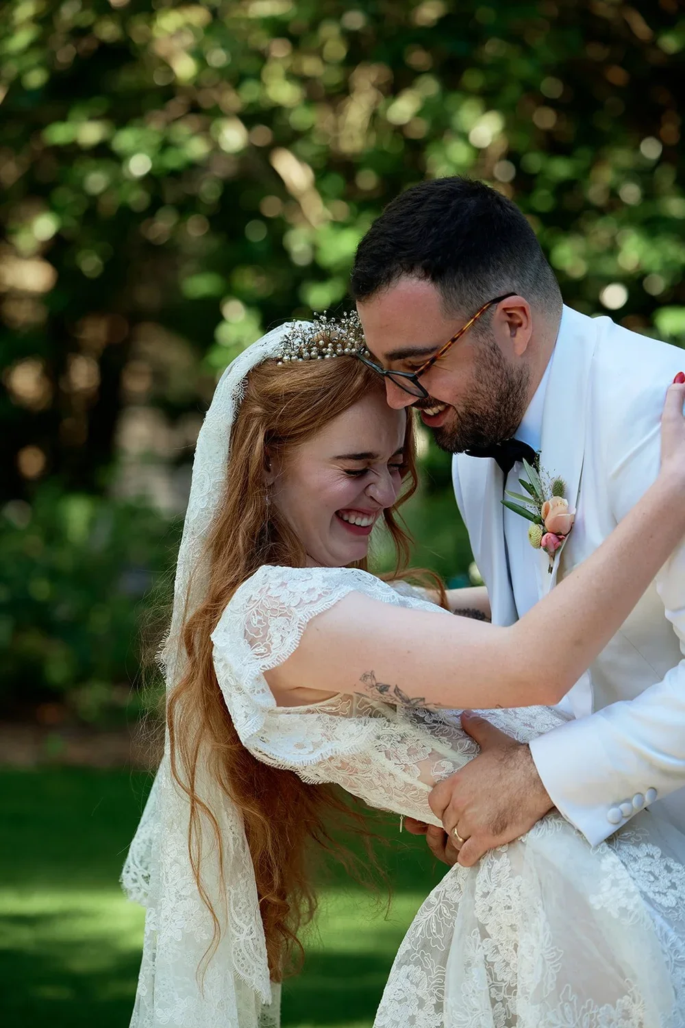 A bride and groom sharing a joyful moment, smiling and leaning their foreheads together, outdoors with greenery in the background.