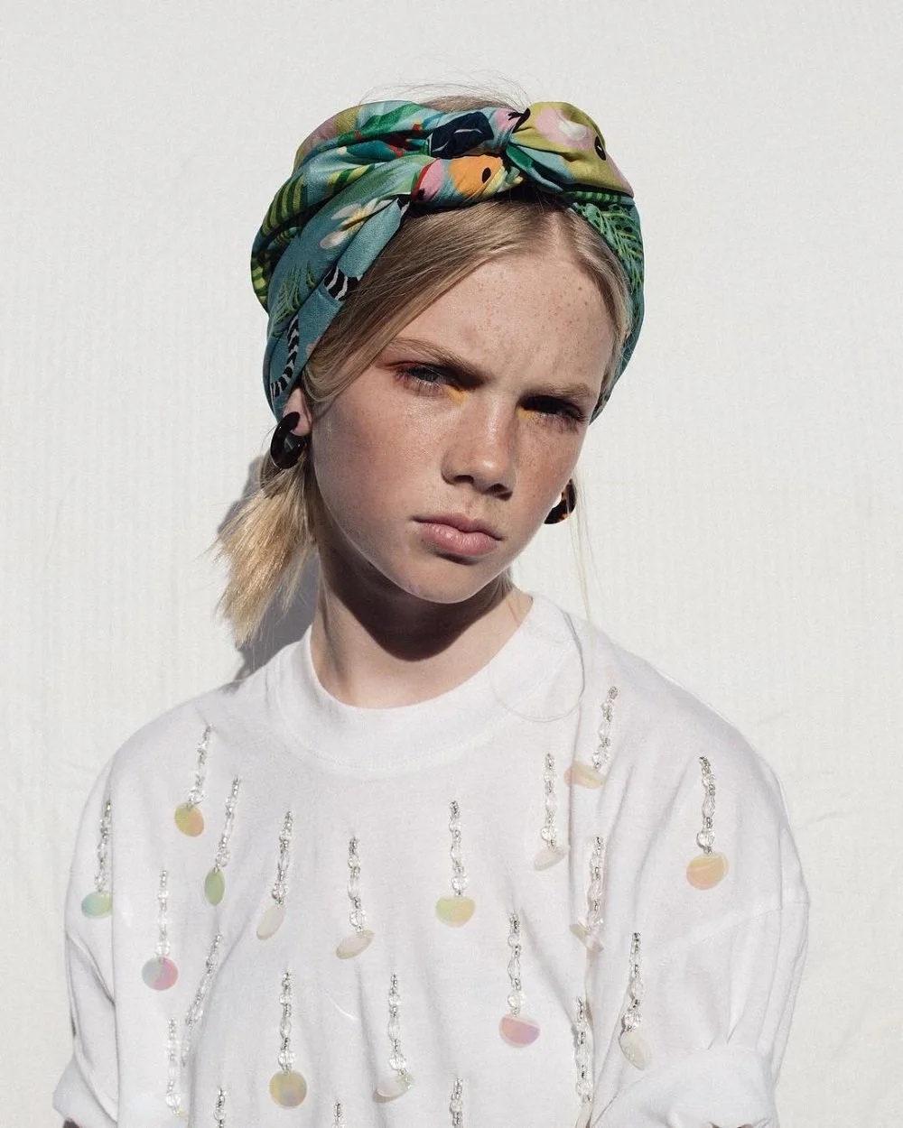 A young woman with blonde hair wearing a colorful headscarf with a pattern of tropical leaves and animals, black earrings, and a white shirt decorated with pastel-colored beads or sequins, standing against a plain white background.
