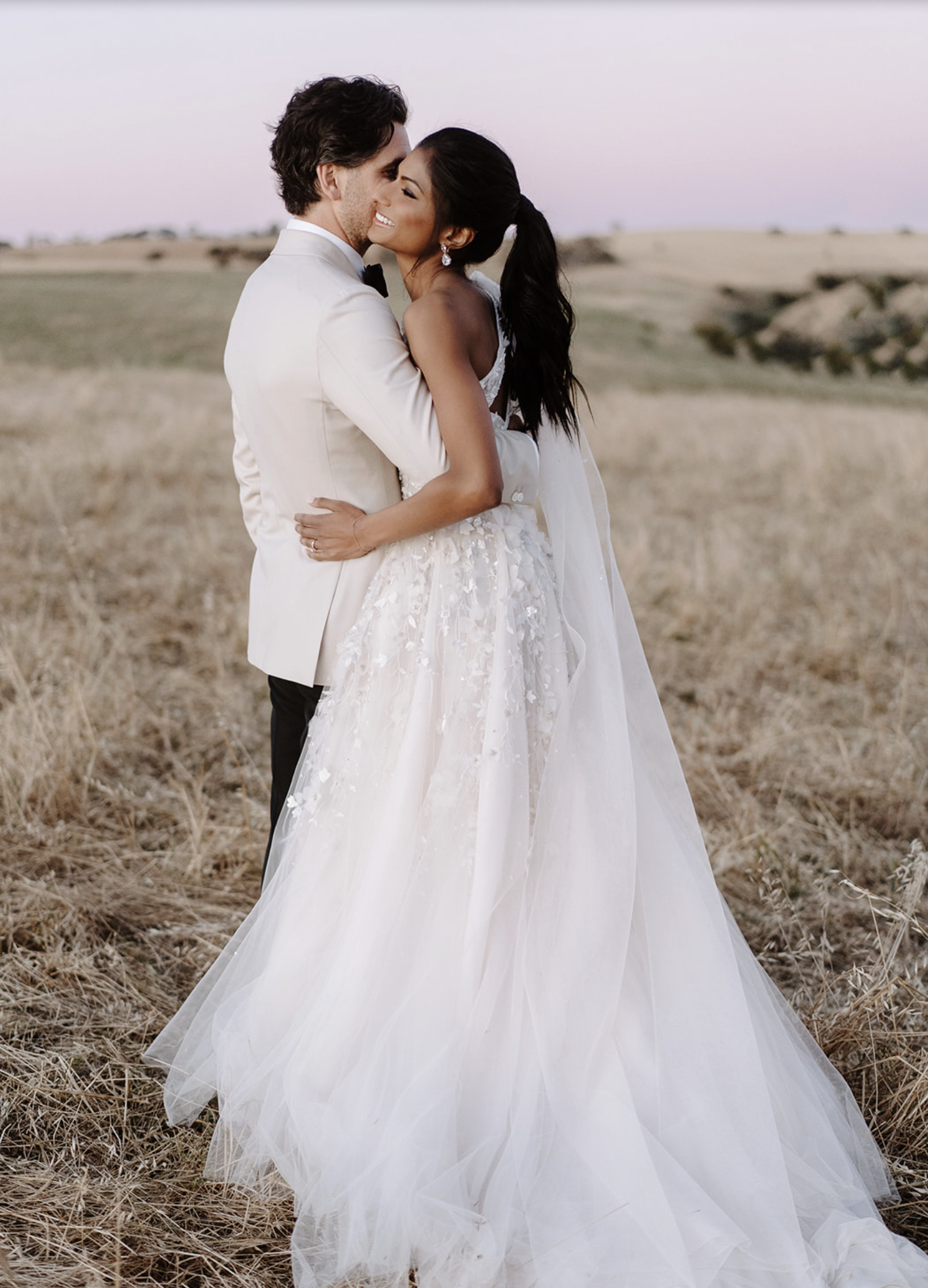 A bride and groom in wedding attire embracing in a field during sunset.