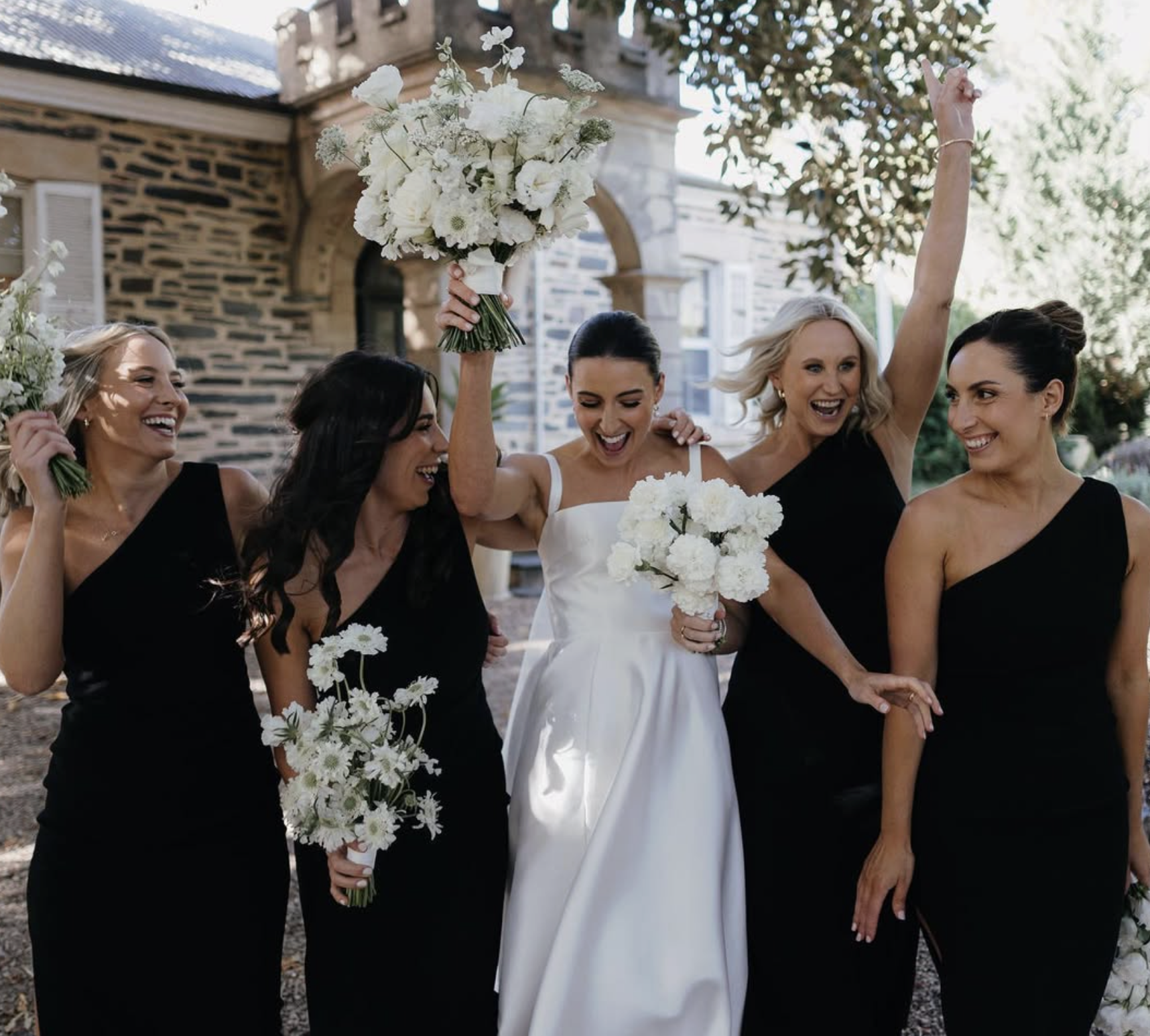 A group of five women celebrating outdoors, dressed in formal attire. The woman in the center is wearing a white wedding dress, holding a bouquet of white flowers, and raising her arm in celebration. The other women are dressed in black, holding bouq