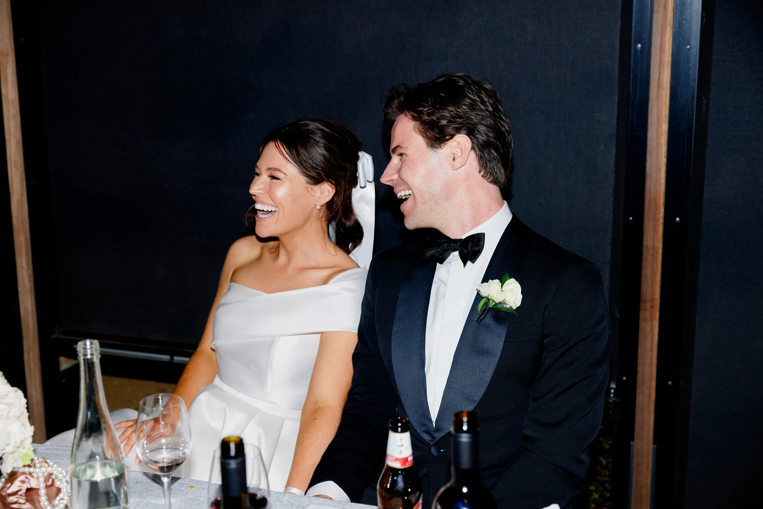 A bride and groom laughing together at their wedding reception, sitting at a table with drinks and a floral centerpiece.